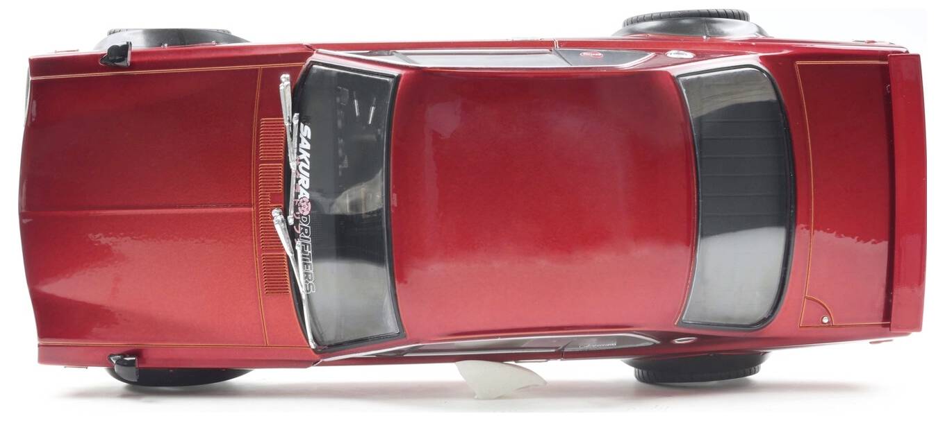 Červený model hračkářského auta pohledem shora, ukazující detailní prvky jako stěrače a boční zrcátka, na jednobarevném bílém pozadí.