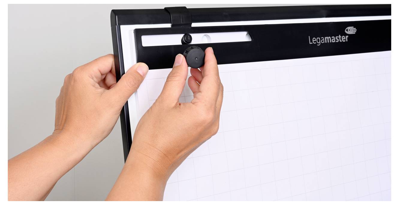 Ruce nastavující ovladač na flipchartu značky Legamaster. Flipchart je připraven k použití a zobrazuje prázdnou mřížku pro psaní nebo prezentace.