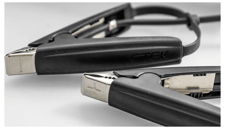 Detailní záběr dvou černých svorek startovacích kabelů na bílém povrchu, zobrazující kovové svorky a kabel propojující je.