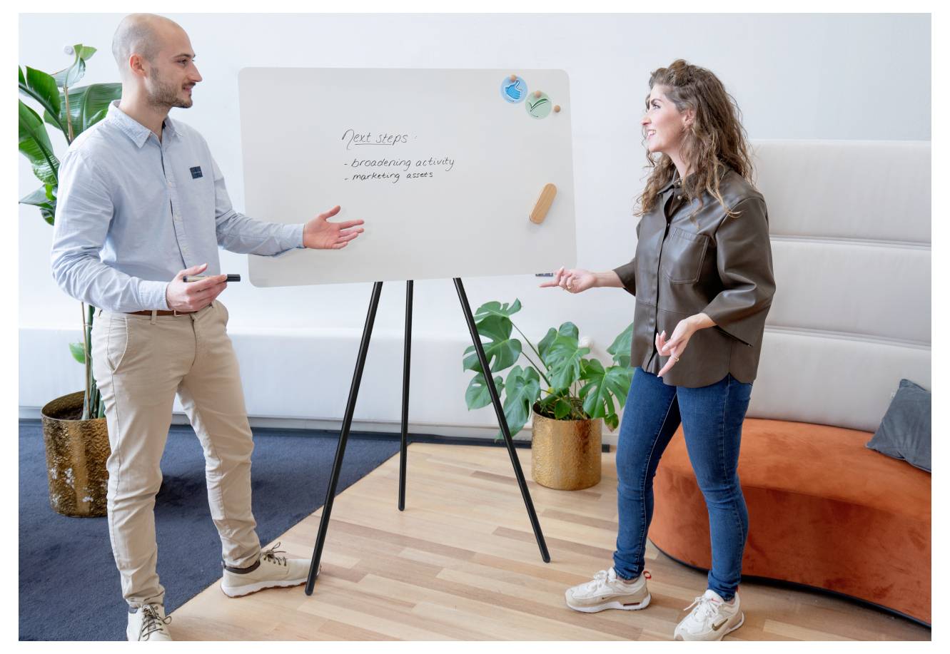 Dva lidé vedou diskusi u whiteboard tabule, kde jsou vypsány 'Další kroky: brainstormingová aktivita, marketingová kontrola' v kancelářském prostředí.