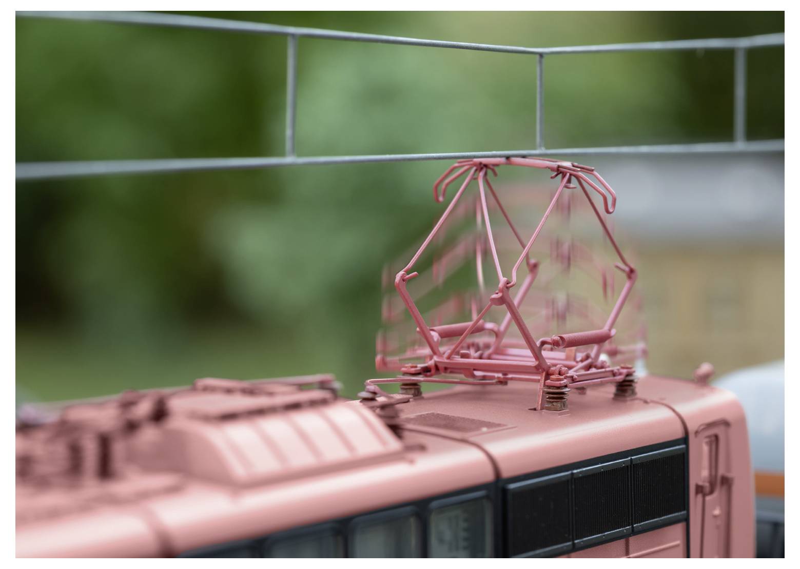 Detail zblízka střechy modelové lokomotivy s komplexním červeným elektrickým zařízením na rozostřeném zeleném pozadí.