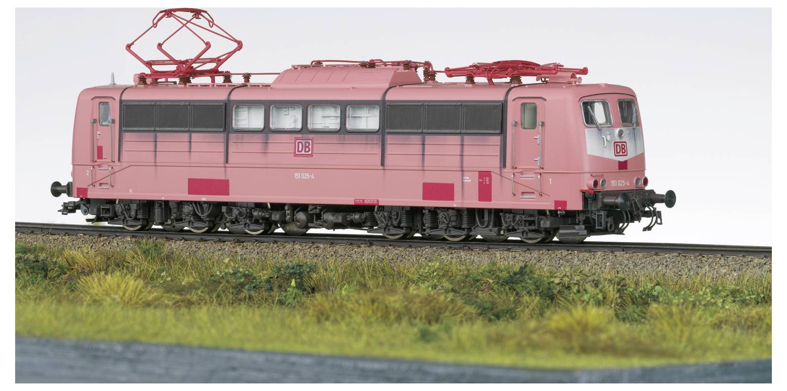 Růžová modelová lokomotiva jedoucí po kolejích s travnatou krajinou. Lokomotiva má na boku červený emblém s bílým písmem.