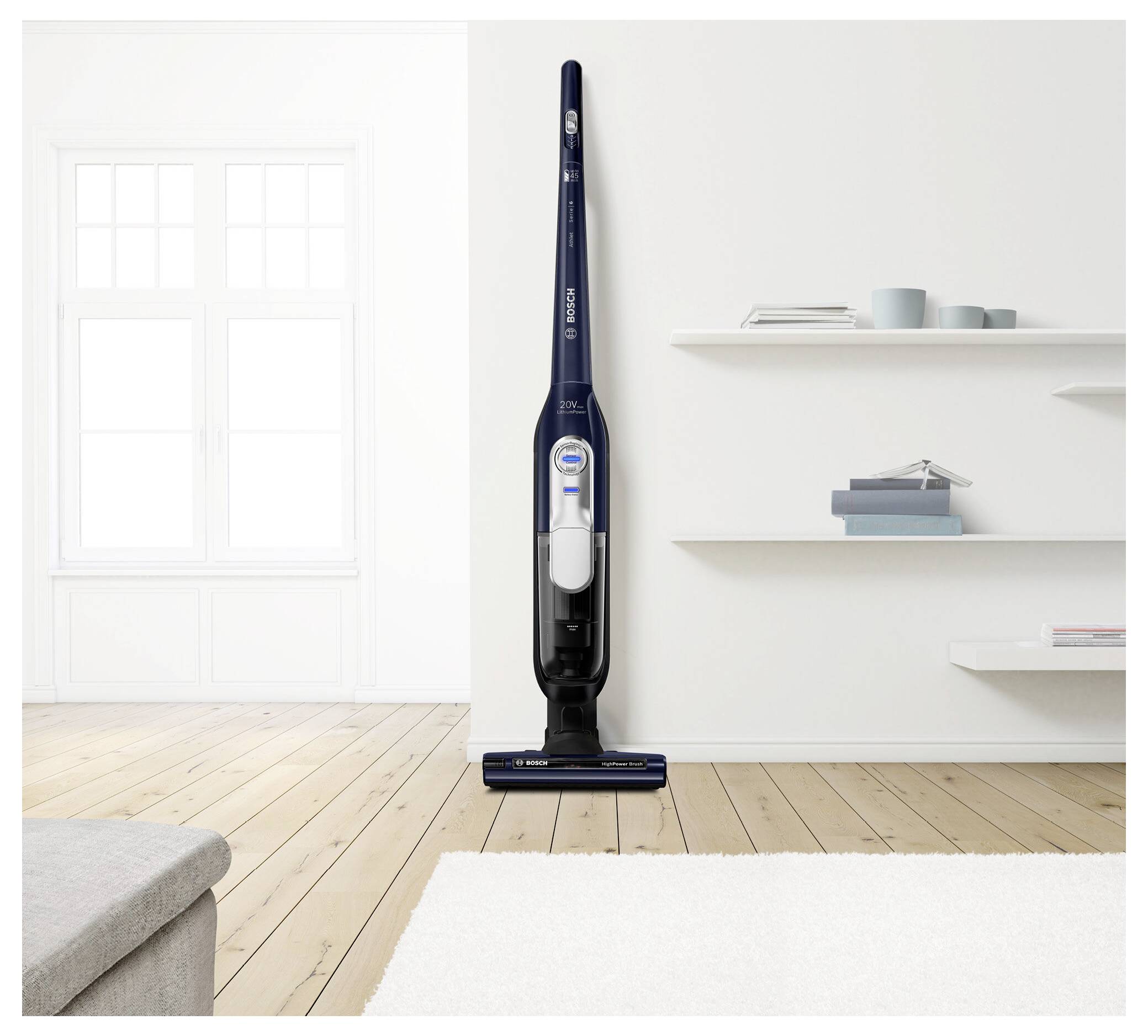 Bezdrátový vysavač stojí vzpřímeně na dřevěné podlaze v moderním, minimalistickém obývacím pokoji s bílými stěnami a kobercem.