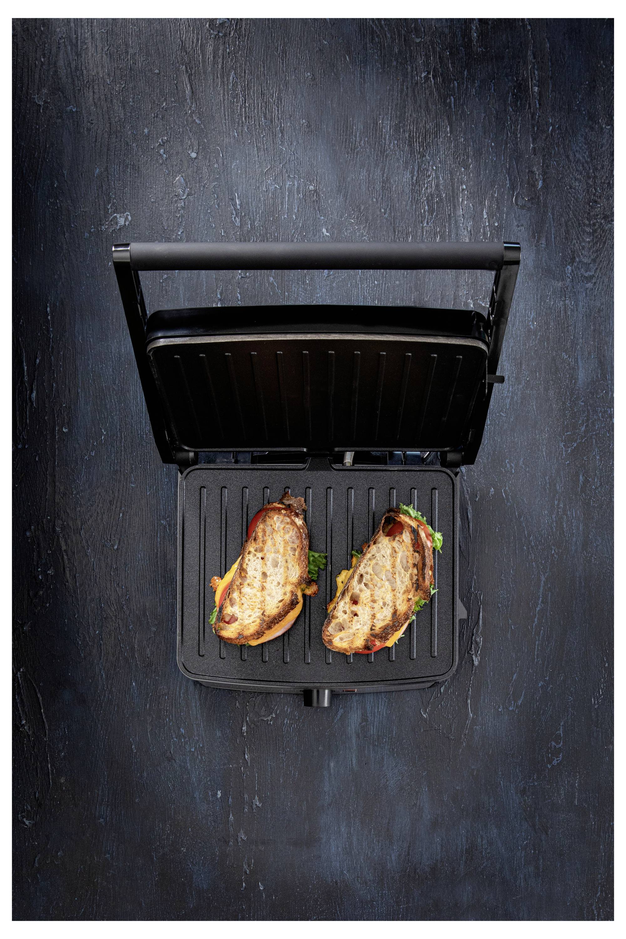 Dva grilované sendviče s rozehřátým sýrem a listem salátu na panini grilu, pohled shora, na strukturovaném tmavém povrchu.
