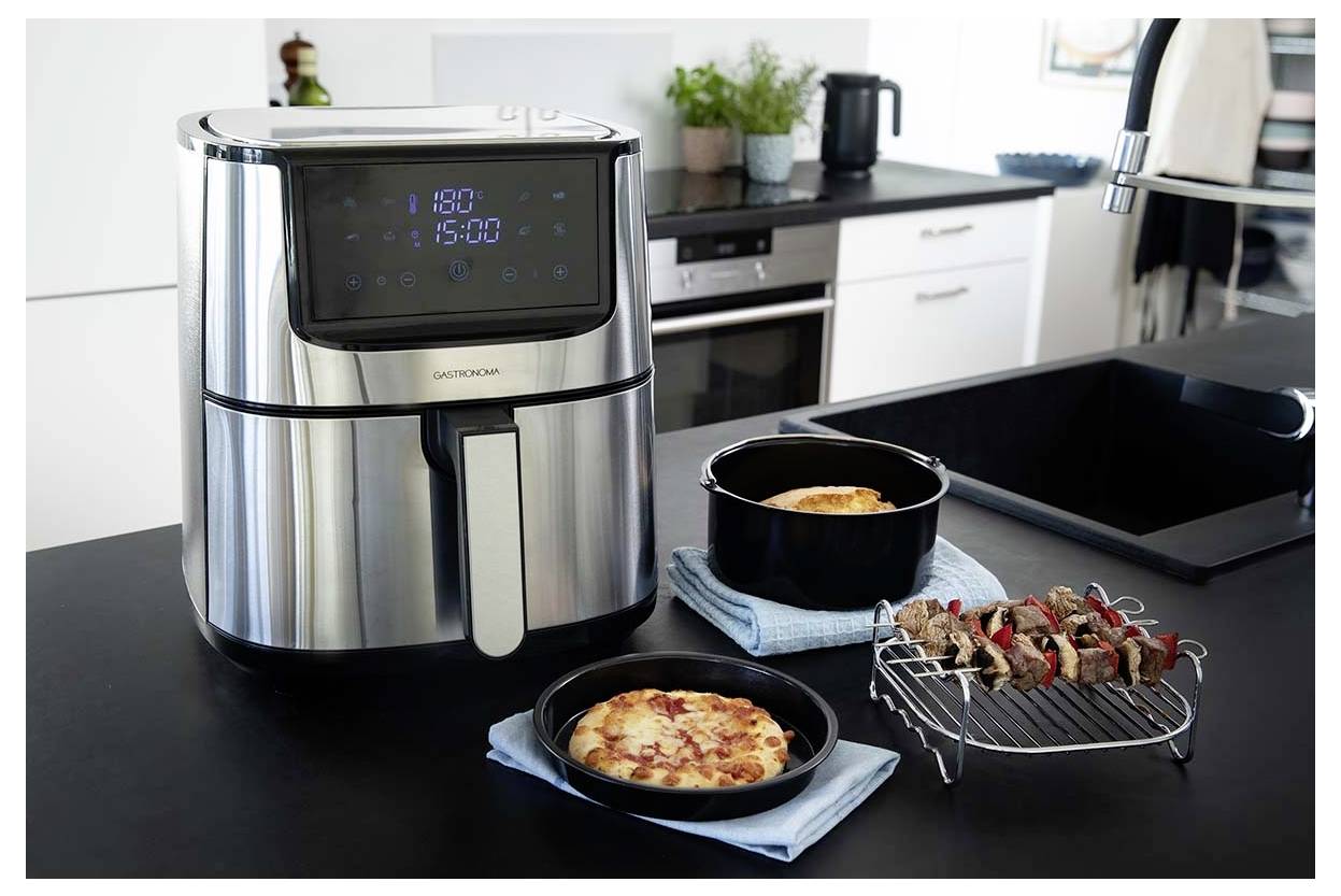 Moderní kuchyně s nerezovou horkovzdušnou fritézou zobrazující '180°' a '15:00'. Poblíž jsou připraveny pizza, zapečený pokrm a špízy.