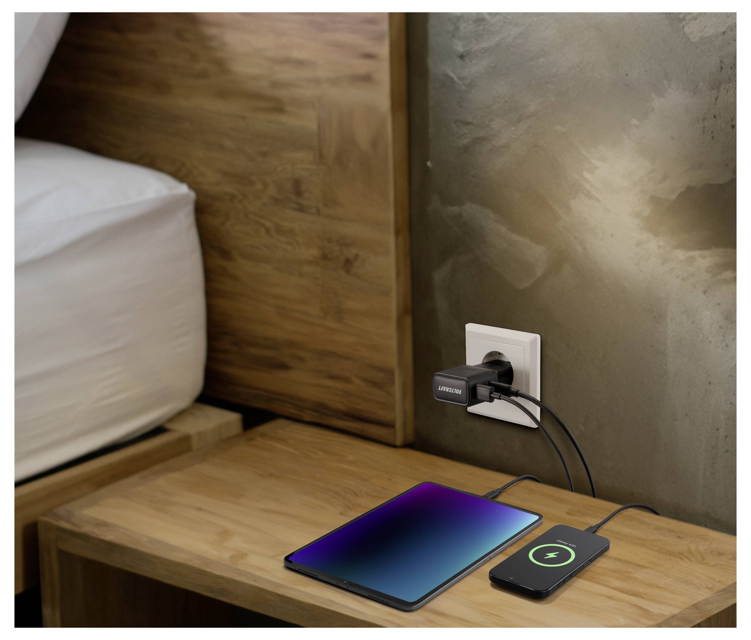 Tablet a smartphone se nabíjejí na dřevěném nočním stolku pomocí duálního USB adaptéru zapojeného do zásuvky ve zdi vedle postele.