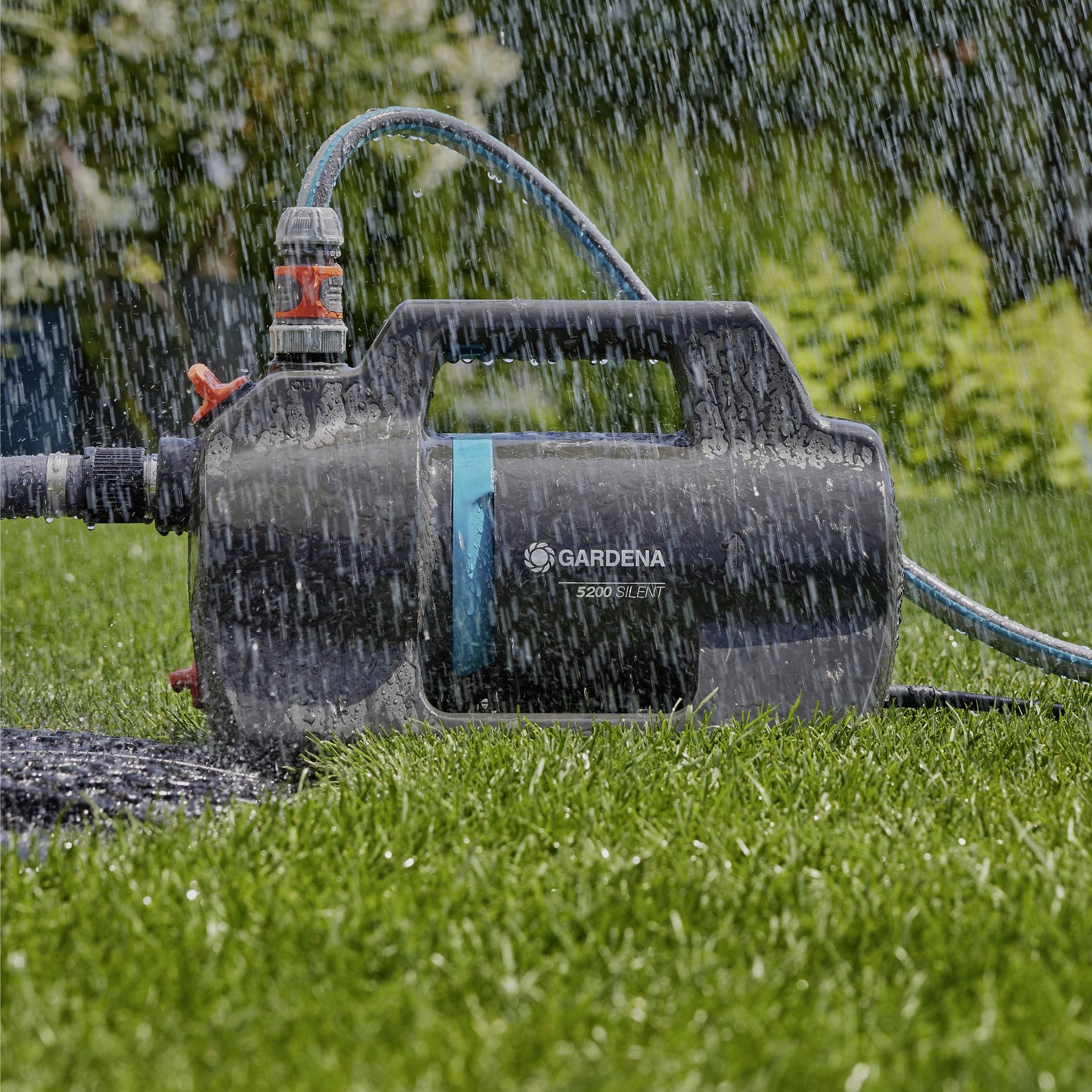 Zahradní čerpadlo označené 'Gardena 3500/4' stojí na mokré trávě, přičemž voda stříká obloukem přes scénu, což naznačuje zavlažování nebo zalévání.