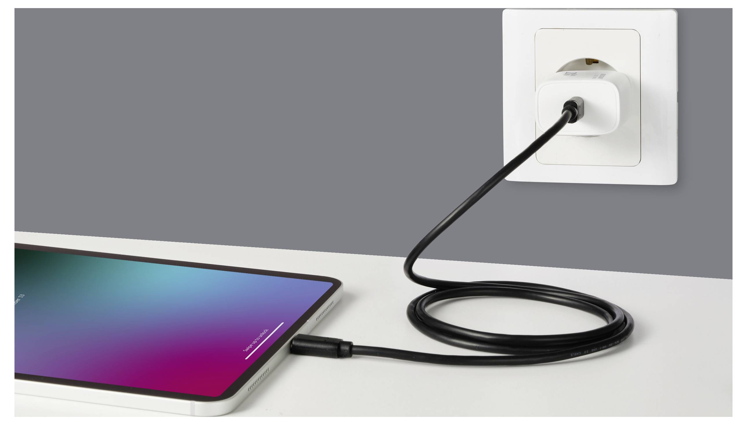 Tablet na stole se nabíjí; černý kabel se připojuje k adaptéru zásuvky na stěně.
