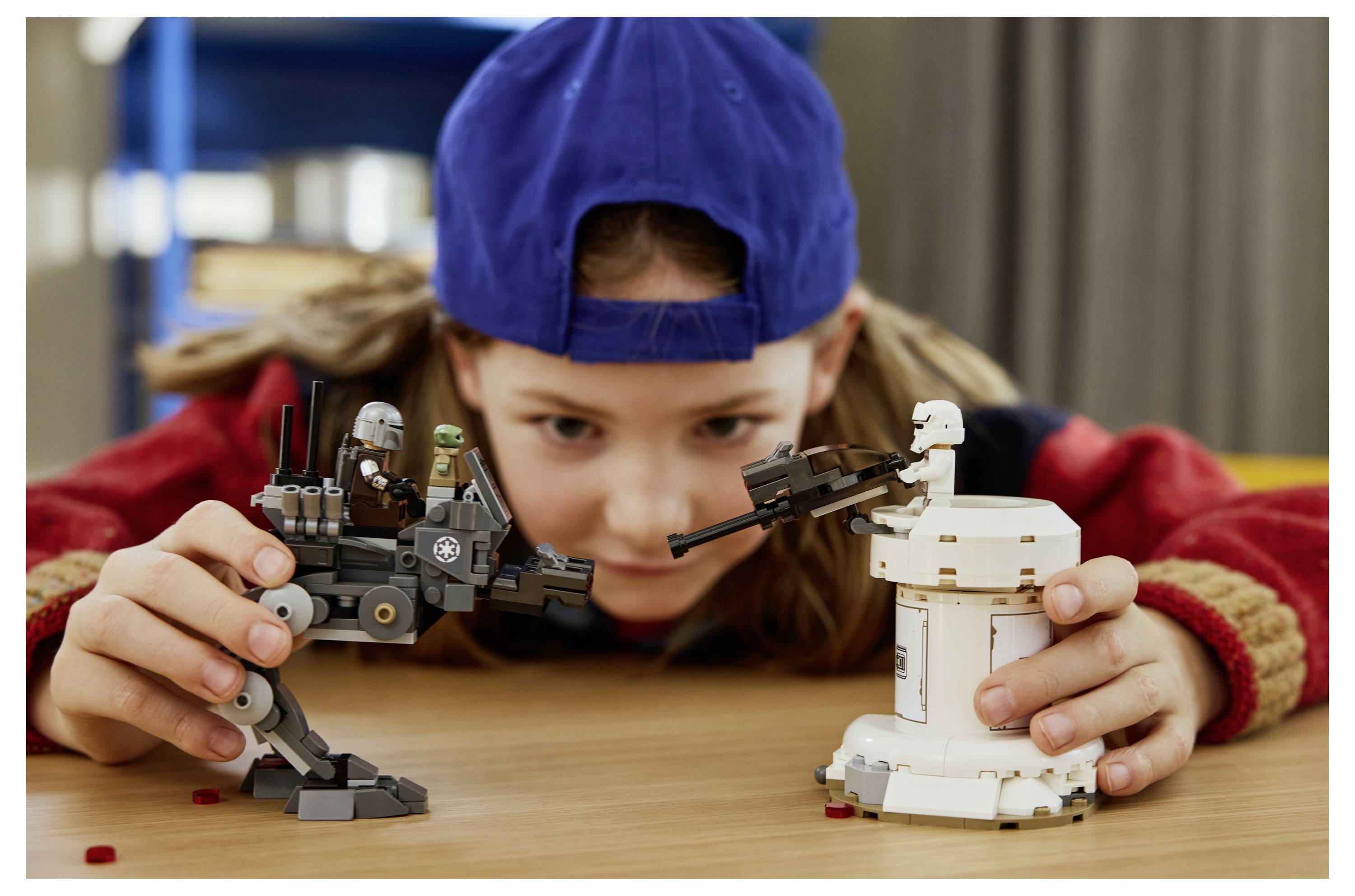 Dítě v modré čepici a červené bundě si hraje s dvěma sadami LEGO s tematikou Star Wars, s šedým chodcem a malou bílou věží, na stole.
