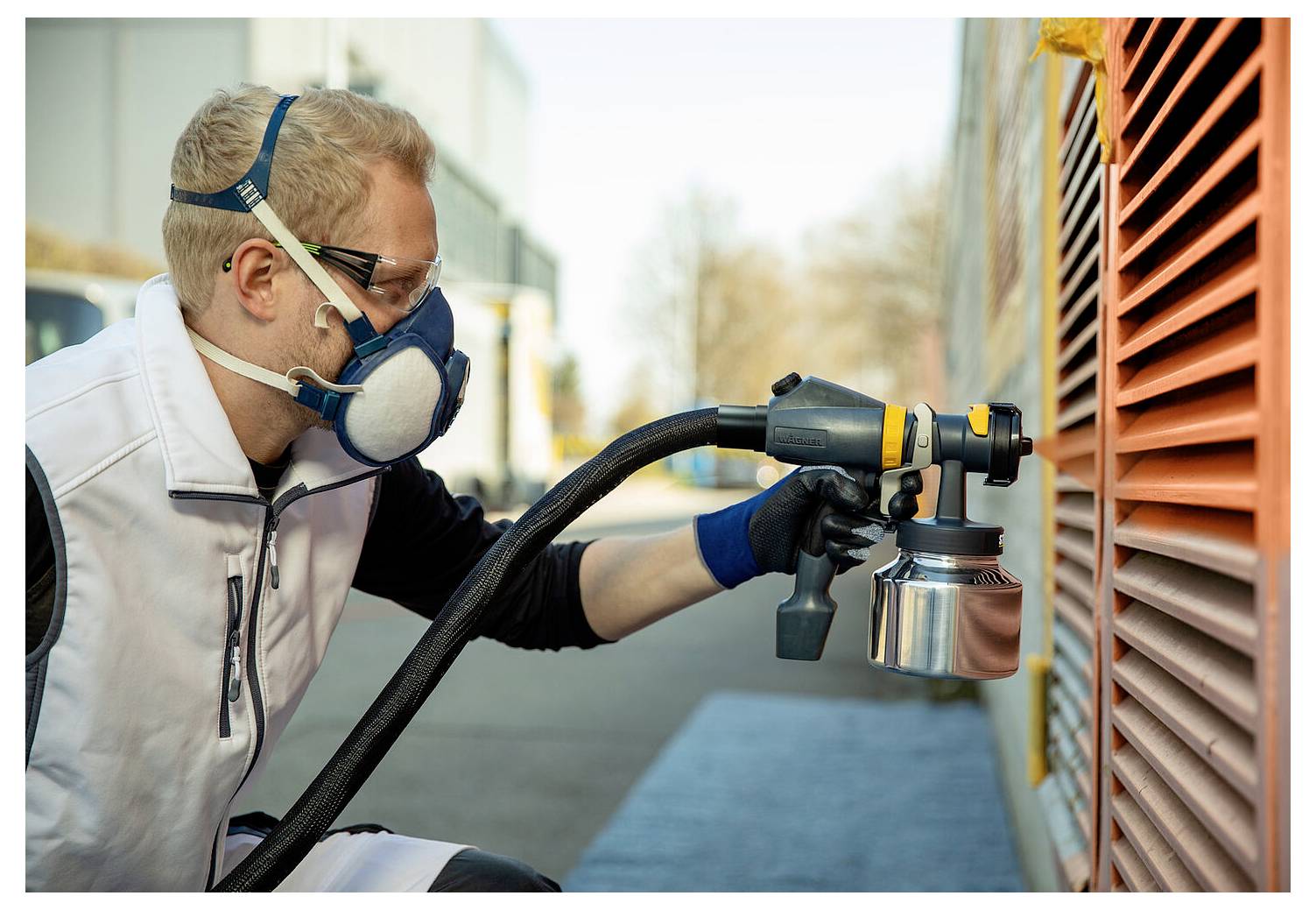 Osoba v ochranném oblečení obsluhuje stříkací pistoli na velkém, zvlněném oranžovém povrchu venku, pravděpodobně jako součást malířské nebo údržbové práce.