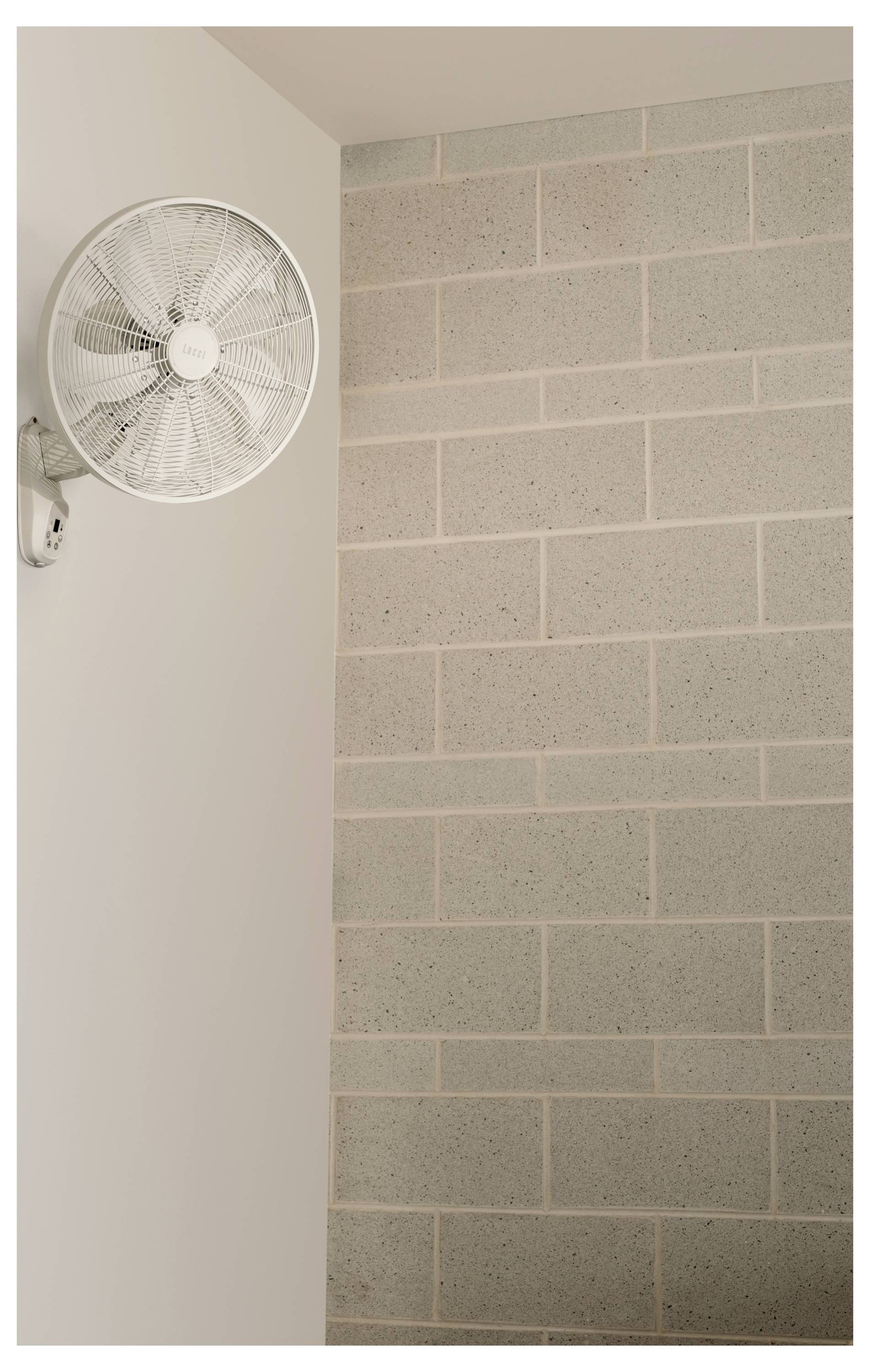 Scéna, kde na stěně visí bílý ventilátor a vedle něj pokračuje zeď z šedých cihel.
