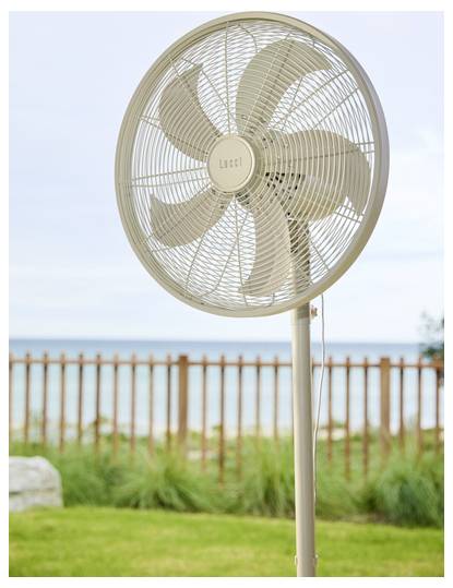 Stojací ventilátor na terase s výhledem na pláž, obklopený dřevěným plotem a trávou, který navozuje atmosféru větrného venkovního prostoru.