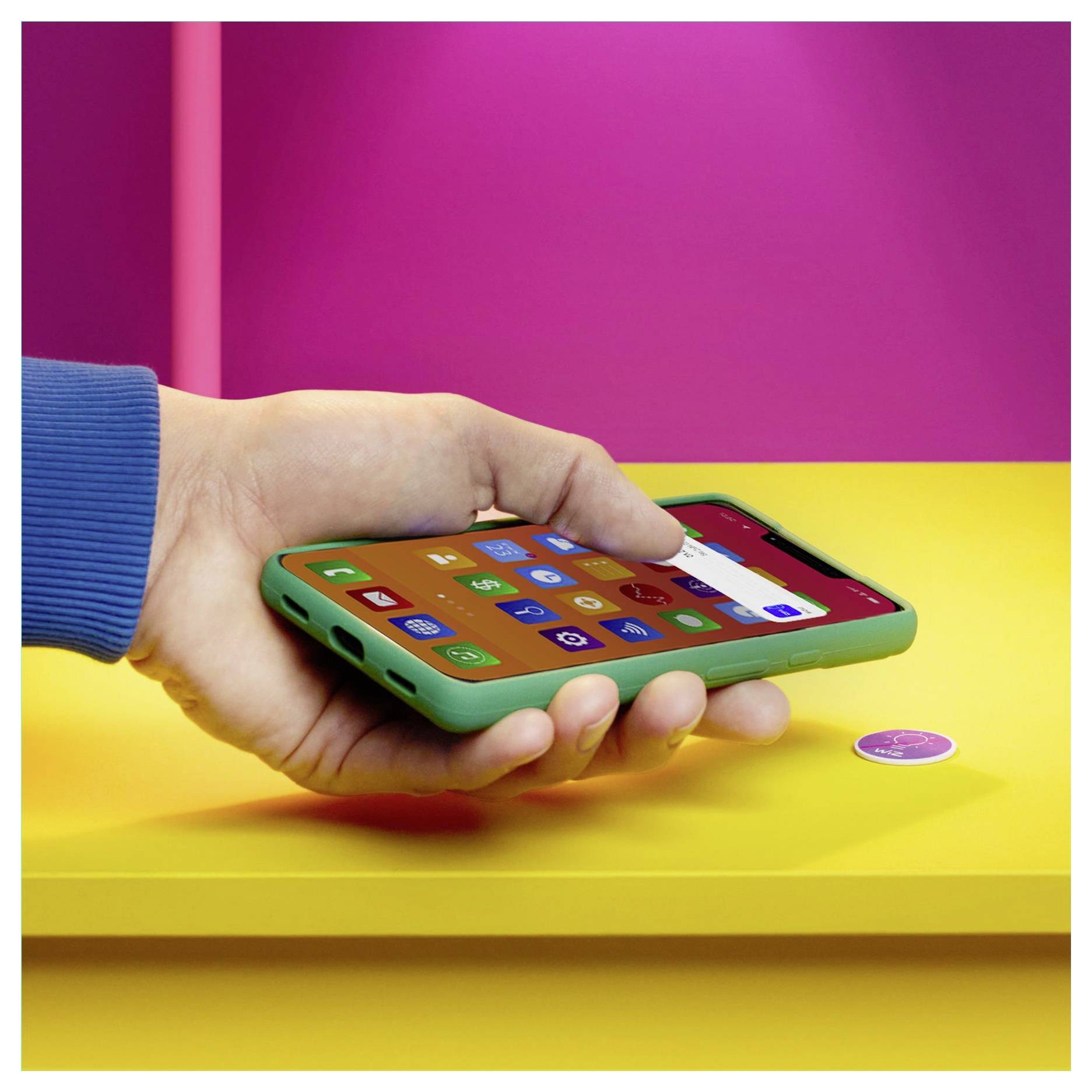 Ruka drží zelený smartphone s barevnými ikonami aplikací viditelnými na displeji. Telefon visí nad žlutým stolem s růžovou stěnou v pozadí.