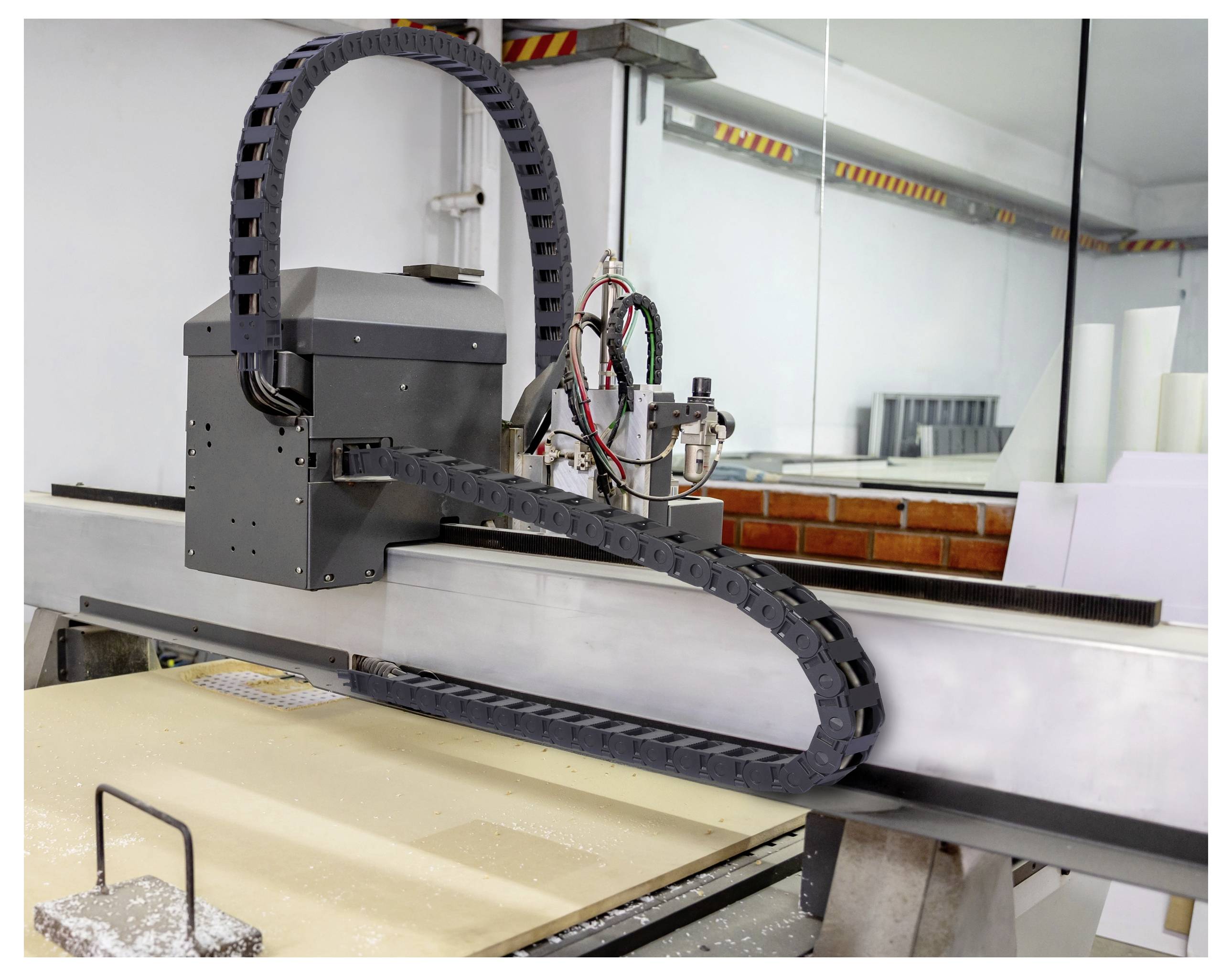 Velký CNC stroj řezující dřevěný panel. Stroj má černou, flexibilní kabelovou dráhu a nachází se v dílně s viditelnými nástroji.
