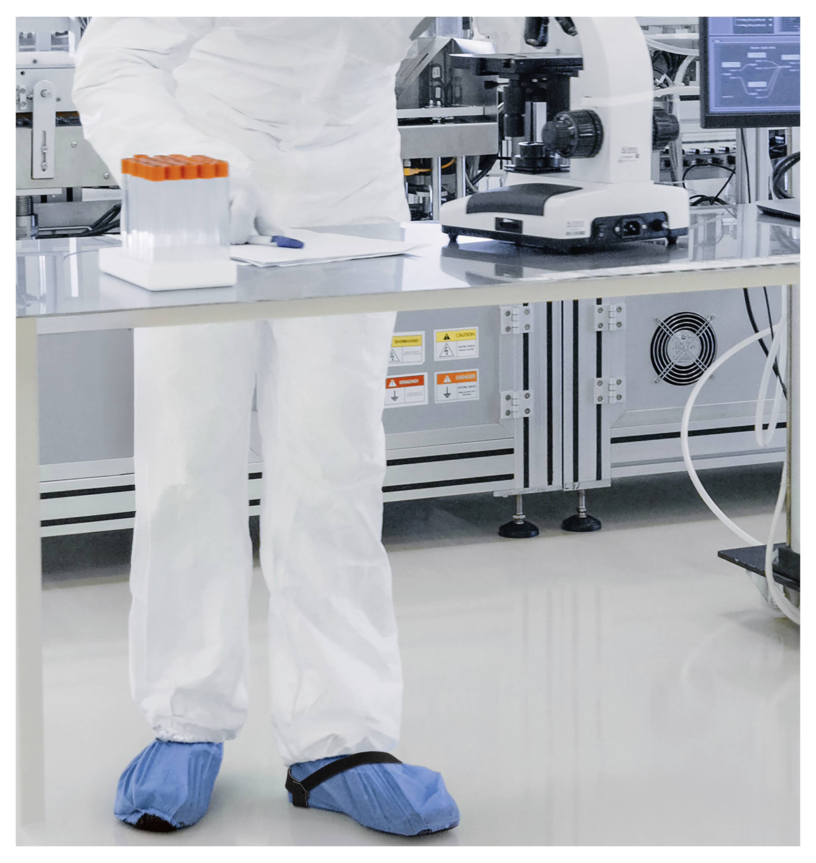 Osoba v oděvu pro čistou místnost a modrých návlecích na boty stojí u laboratorního stolu s epruvetami a používá mikroskop.