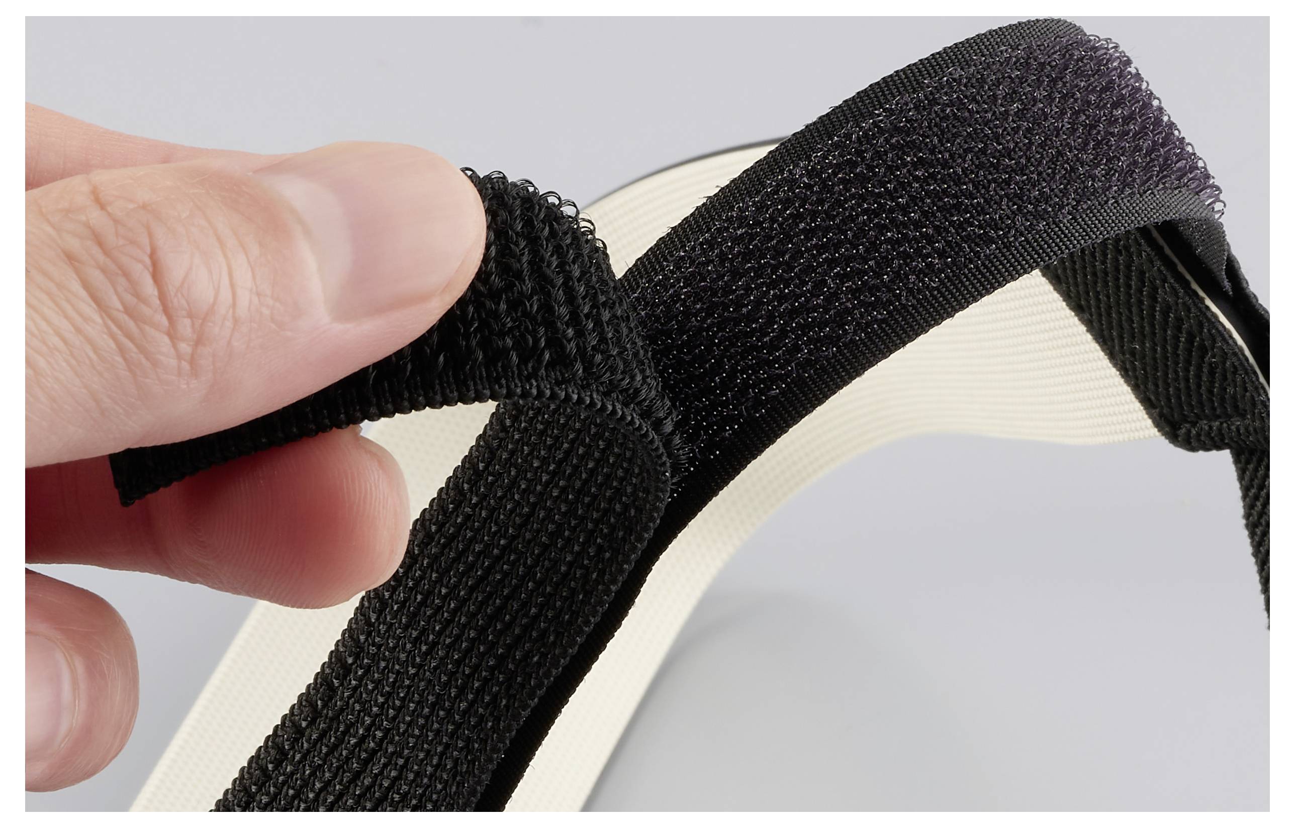 Ruka upravuje černý Velcro pás na lehce šedém pozadí, což demonstruje, jak jej zajistit nebo uvolnit.