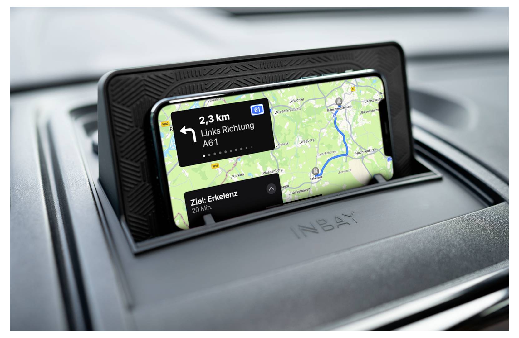 Smartphone zobrazený v přihrádce automobilového palubního desky ukazující navádění trasy po jednotlivých zatáčkách s modrou trasou a pokyny směrem na 'A61' a 'Cíl: Erkelenz'.