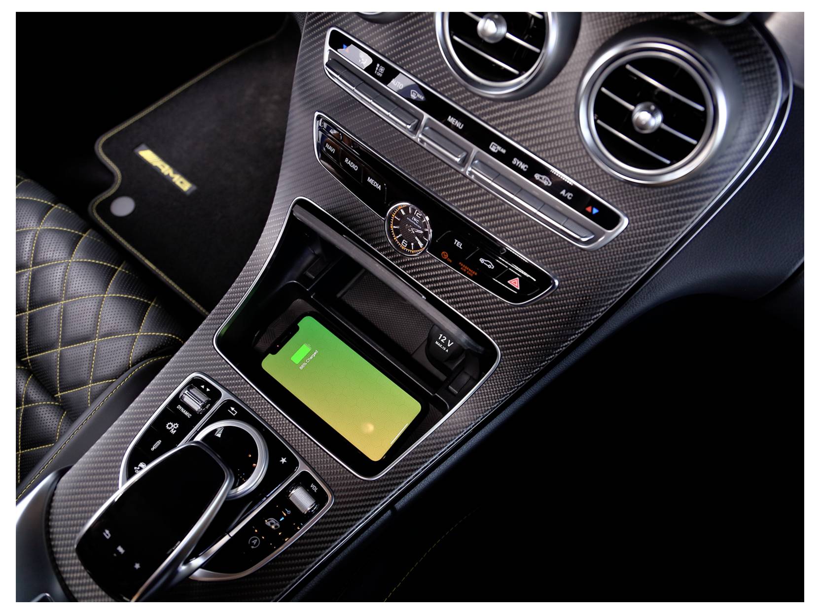 Středový panel automobilu s bezdrátovou nabíjecí podložkou, ovládacími prvky klimatizace, hodinama, odvětráváním a výběrem převodů; viditelný zelený indikátor nabíjení telefonu.