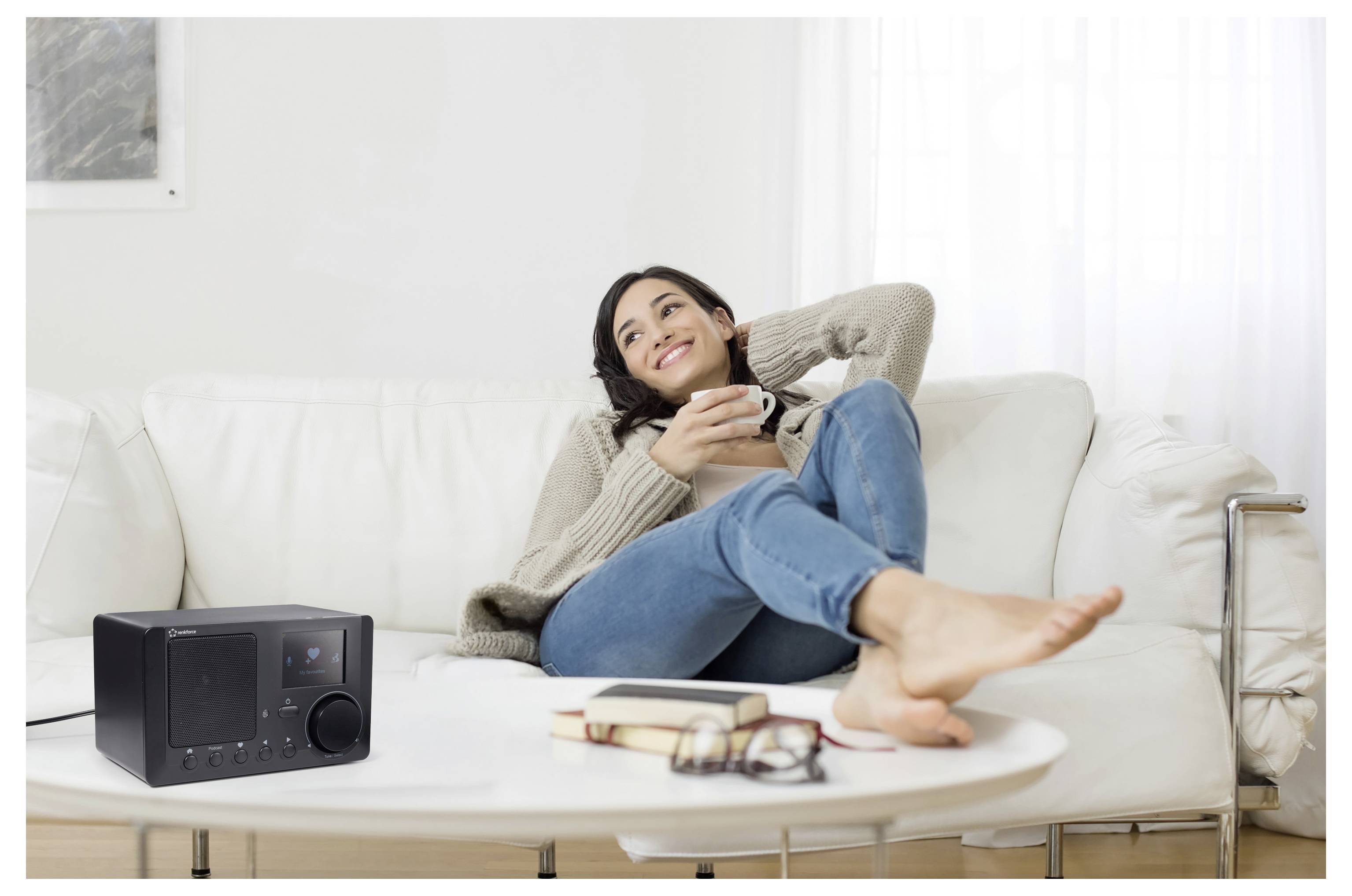 Žena relaxující na bílé pohovce, usmívající se při držení hrnku; na stole vedle knih a brýlí sedí rádio.