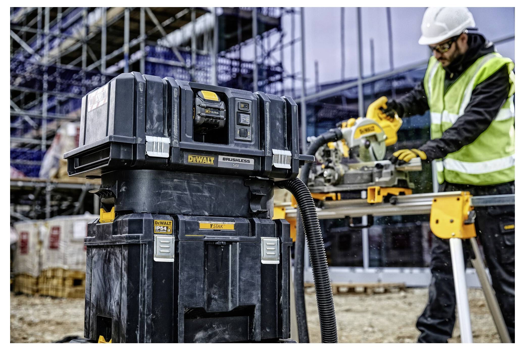 Stavbyhlední pracovník používající stolovací pilu DeWalt na staveništi, se lešením a extraktor prachu DEWALT DWV902M v pozadí.