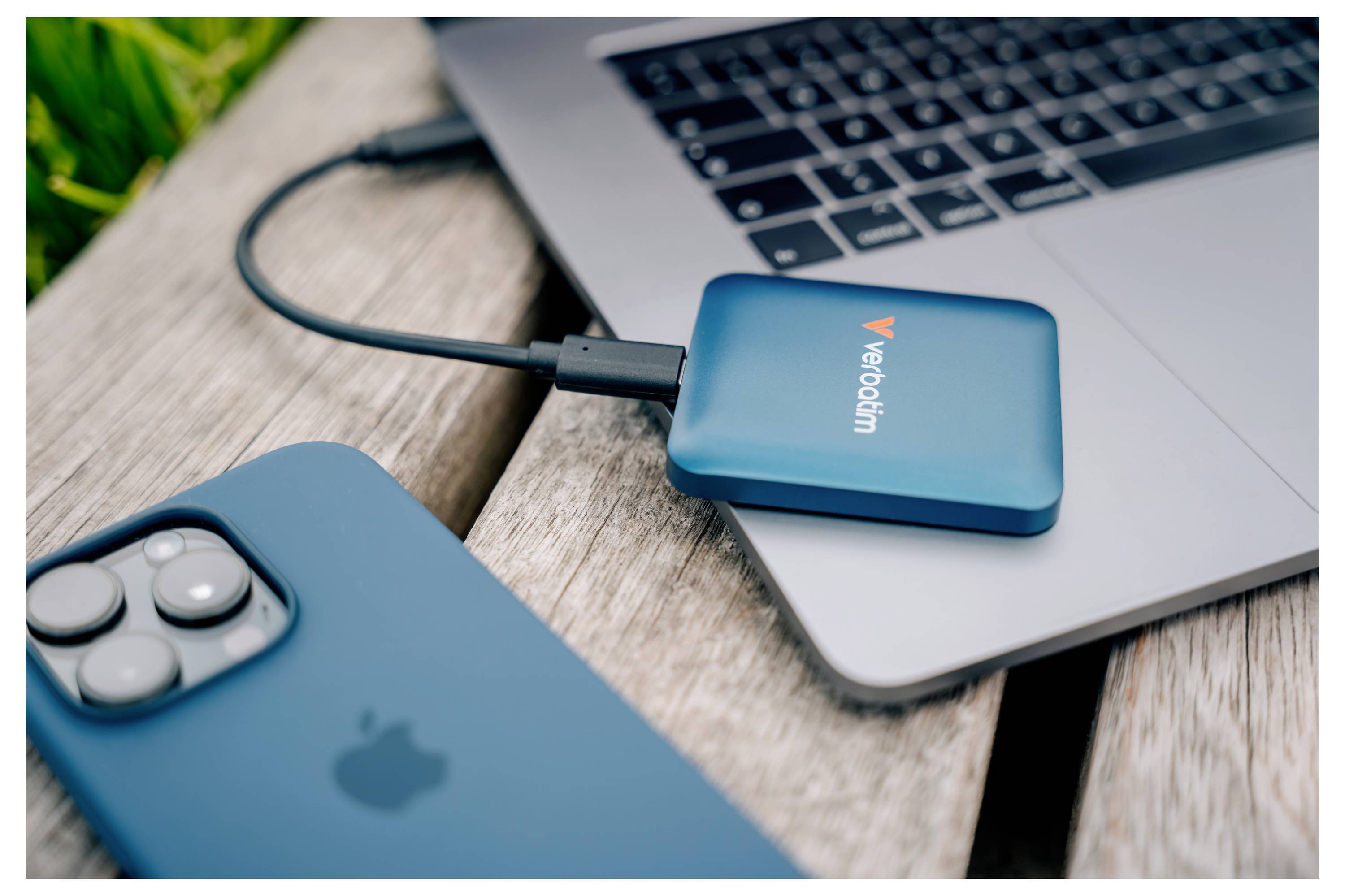 Modrá externí SSD Verbatim připojená přes USB-C k notebooku na dřevěném stole s modrým iPhonem v popředí.
