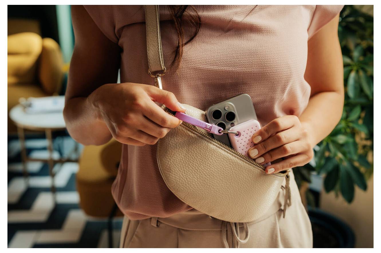 Žena držící růžový zubní kartáček a chytrý telefon v béžové crossbody tašce, která naznačuje cestovní nebo denní nezbytnosti.