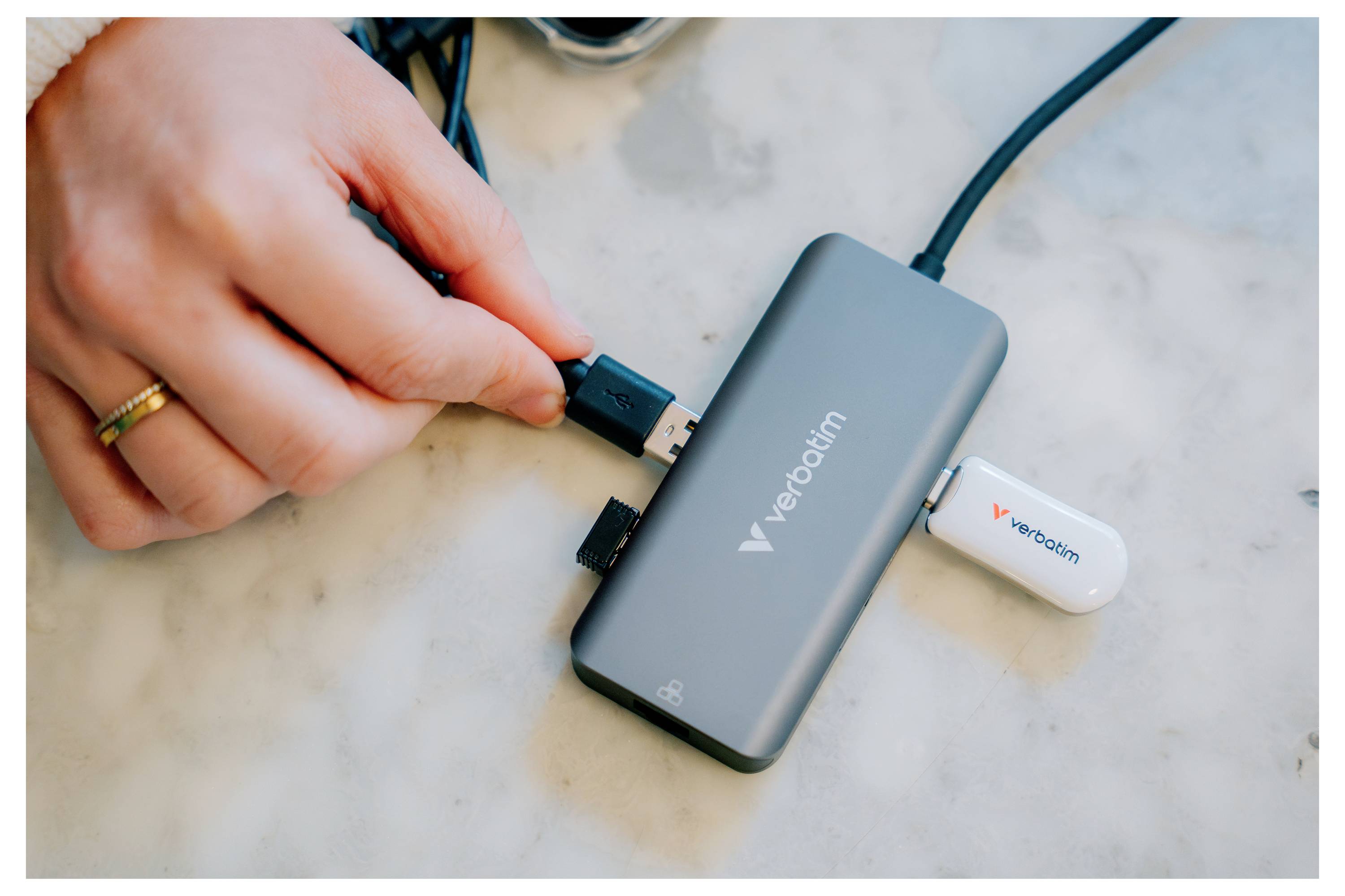 Ruka zapojuje kabel USB do šedého hubu Verbatim s bílou USB flash pamětí připojenou na mramorovém povrchu.
