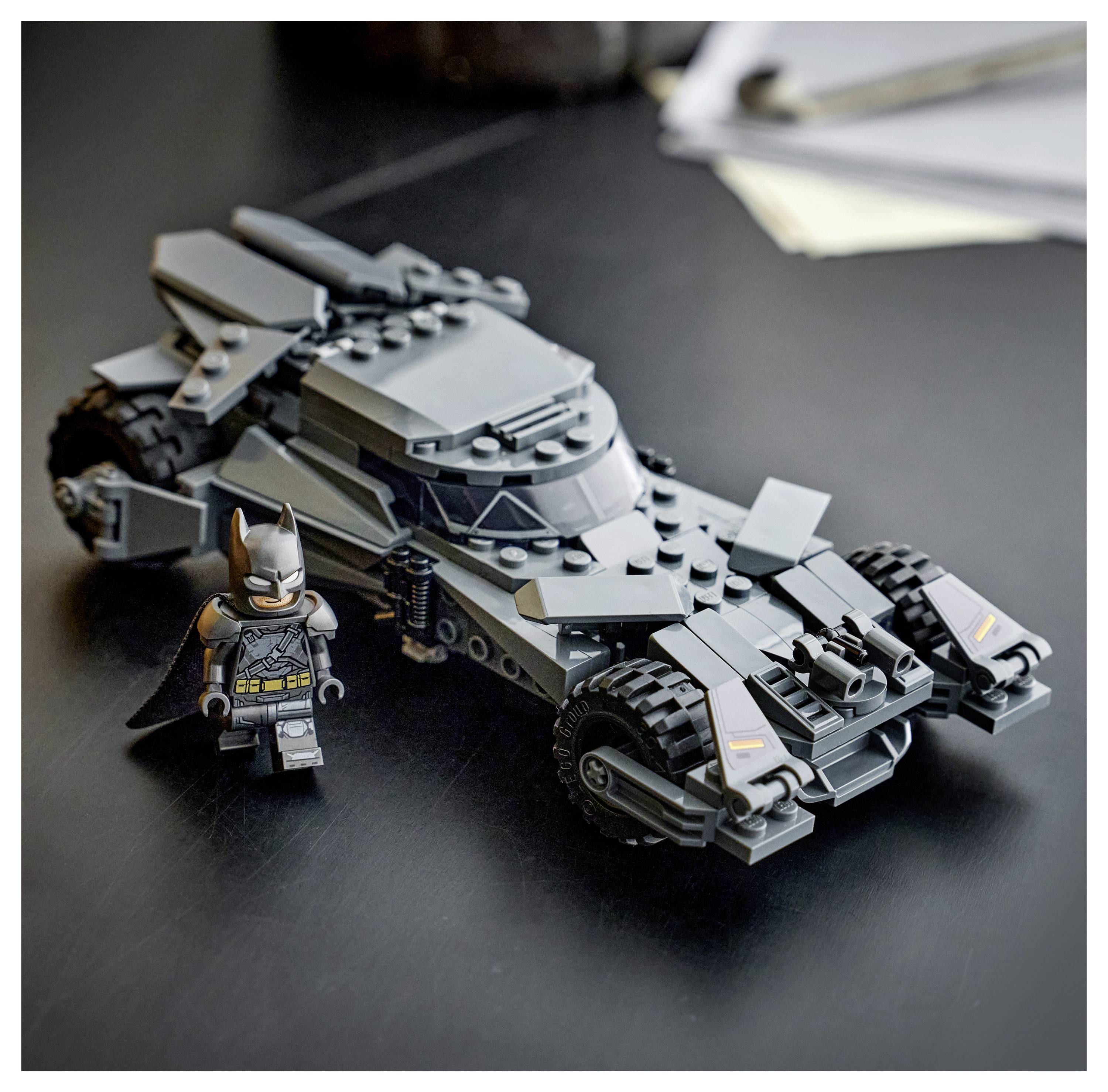 LEGO model černého Batmobilu s figurkou Batmana, umístěný na tmavém stole poblíž papírů. Batmobil má ostré designové prvky.