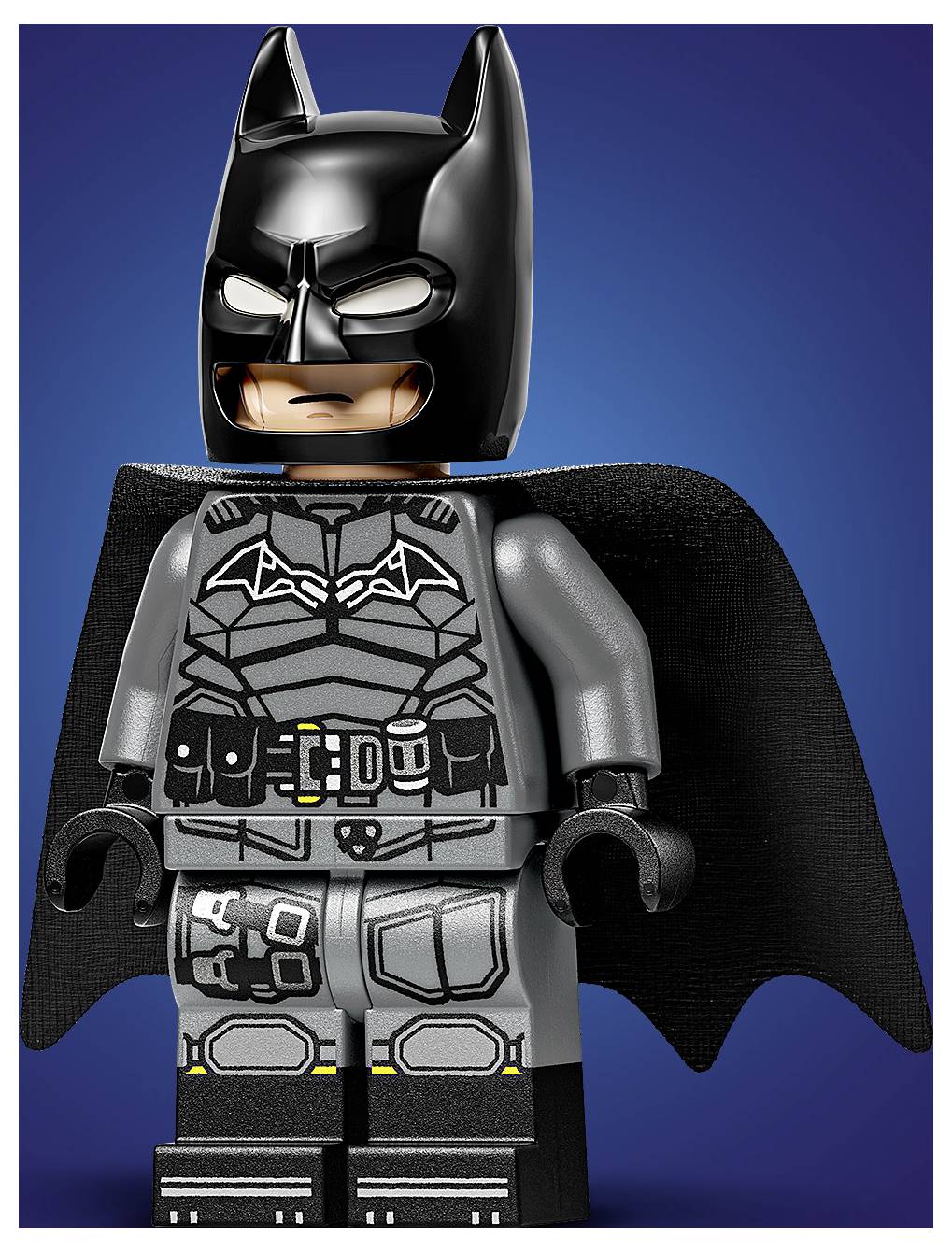 Figurka LEGO superhrdinů v černém obléku, s emblémem netopýra na hrudi a pláštěm, stojící před modrým pozadím.