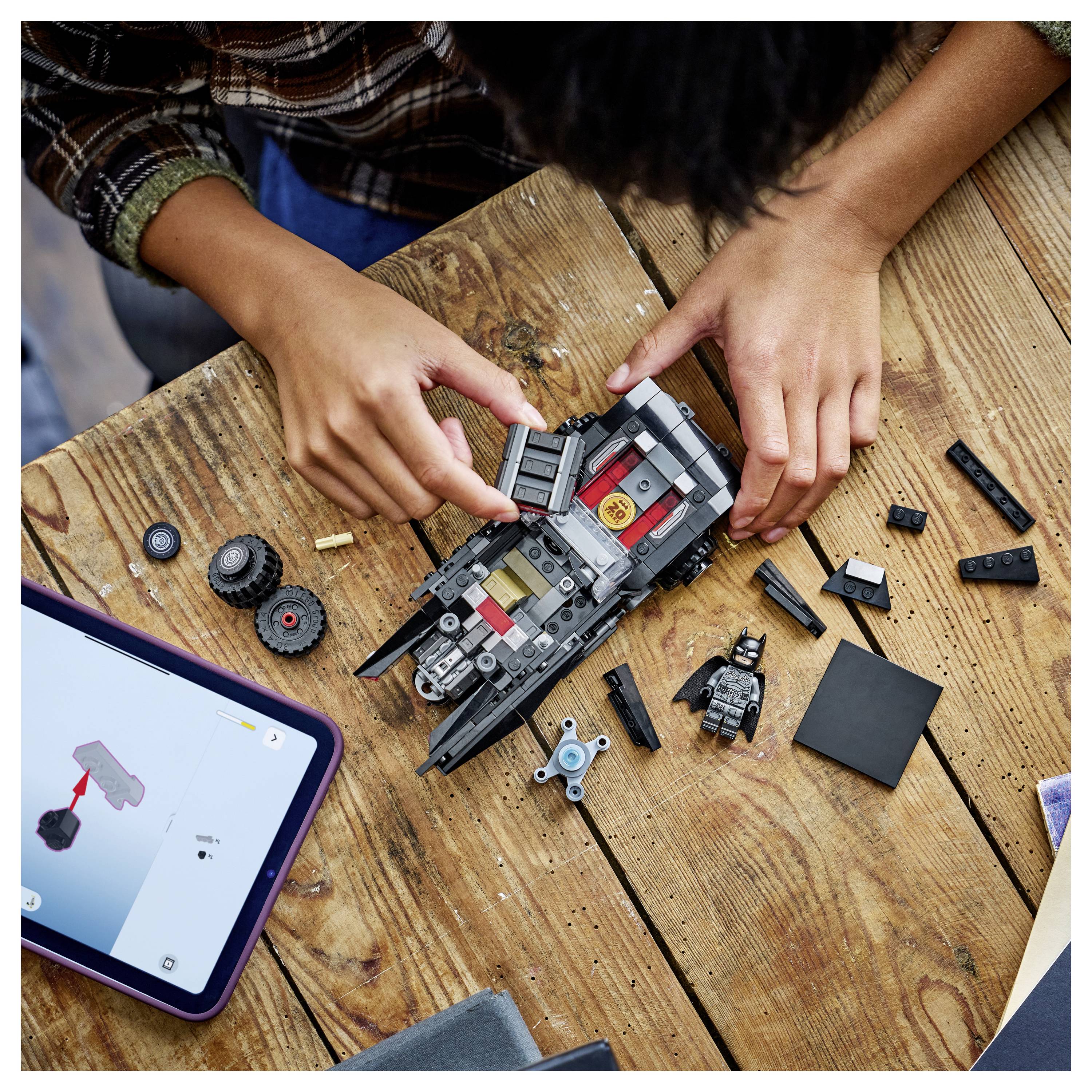 Osoba skládá LEGO Batmobil na dřevěném stole s pokyny zobrazenými na tabletu a různými LEGO kostičkami rozptýlenými kolem.