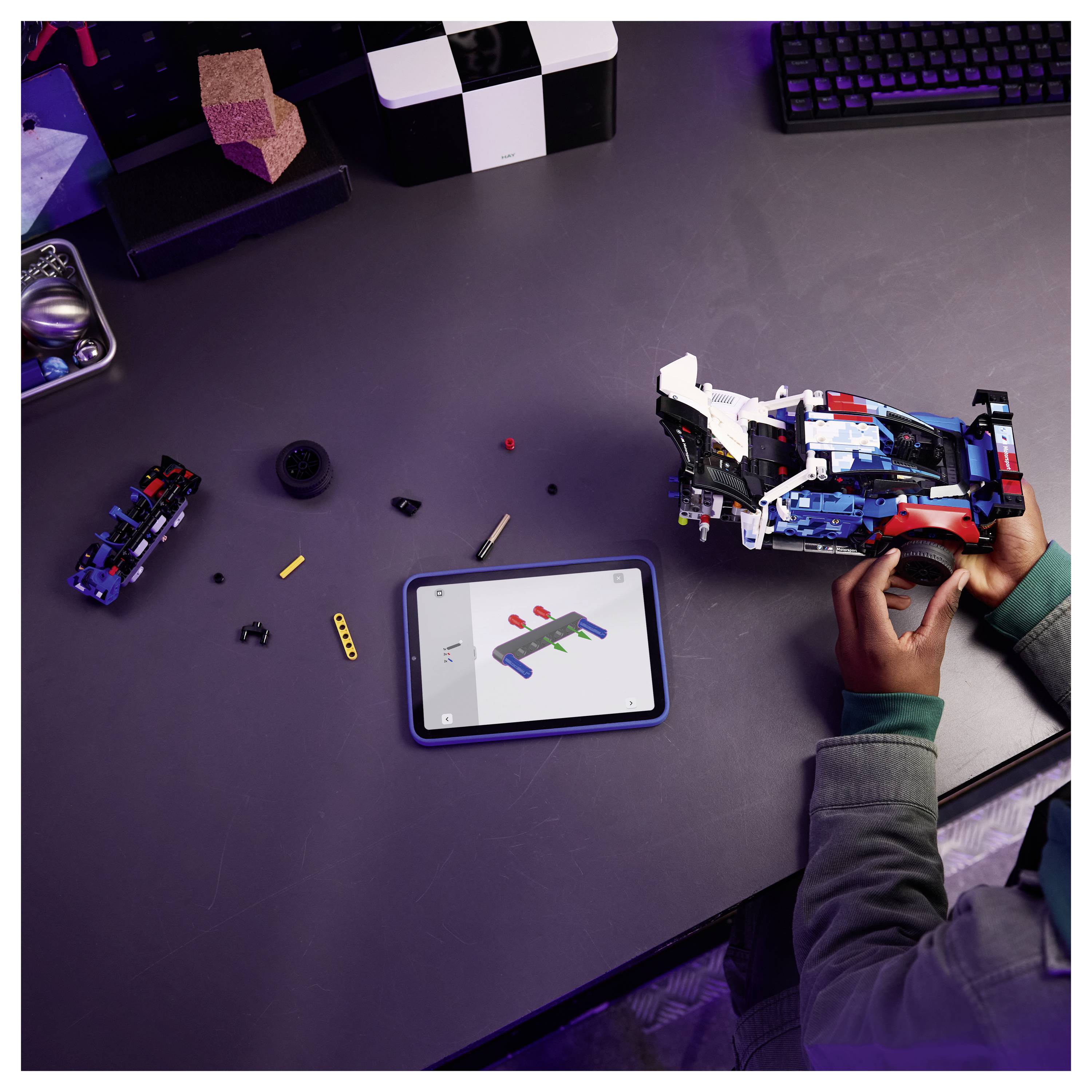 Osoba skládající model LEGO s tabletem zobrazujícím pokyny. Na stole jsou rozloženy různé kusy LEGO a nástroje.