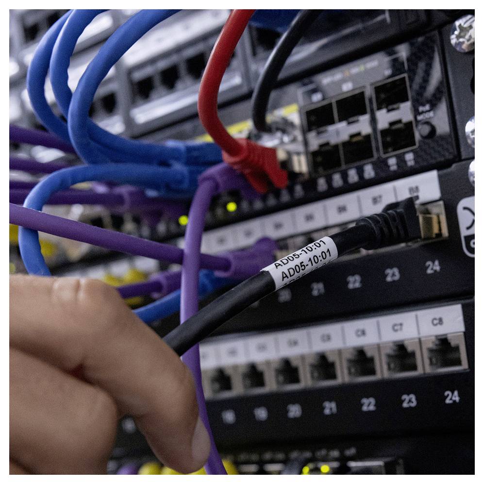 Osoba připojuje černý síťový kabel označený 'AD05-10-01' k serverovému racku plnému kabelů různých barev a přepínačů.