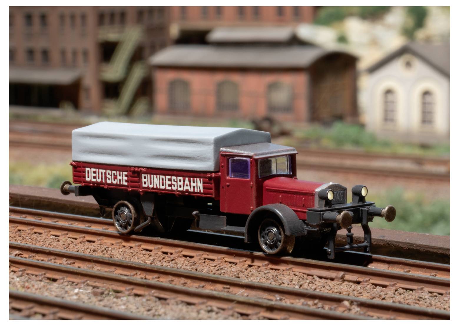 Modelový vlak s nápisem "Deutsche Bundesbahn" na boku stojí na železničních kolejích s pozadím průmyslově stylizovaných budov.