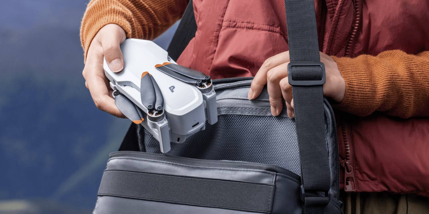 Osoba vkládající kompaktní dron do šedé tašky; oblečená v červené bundě a oranžovém svetru.