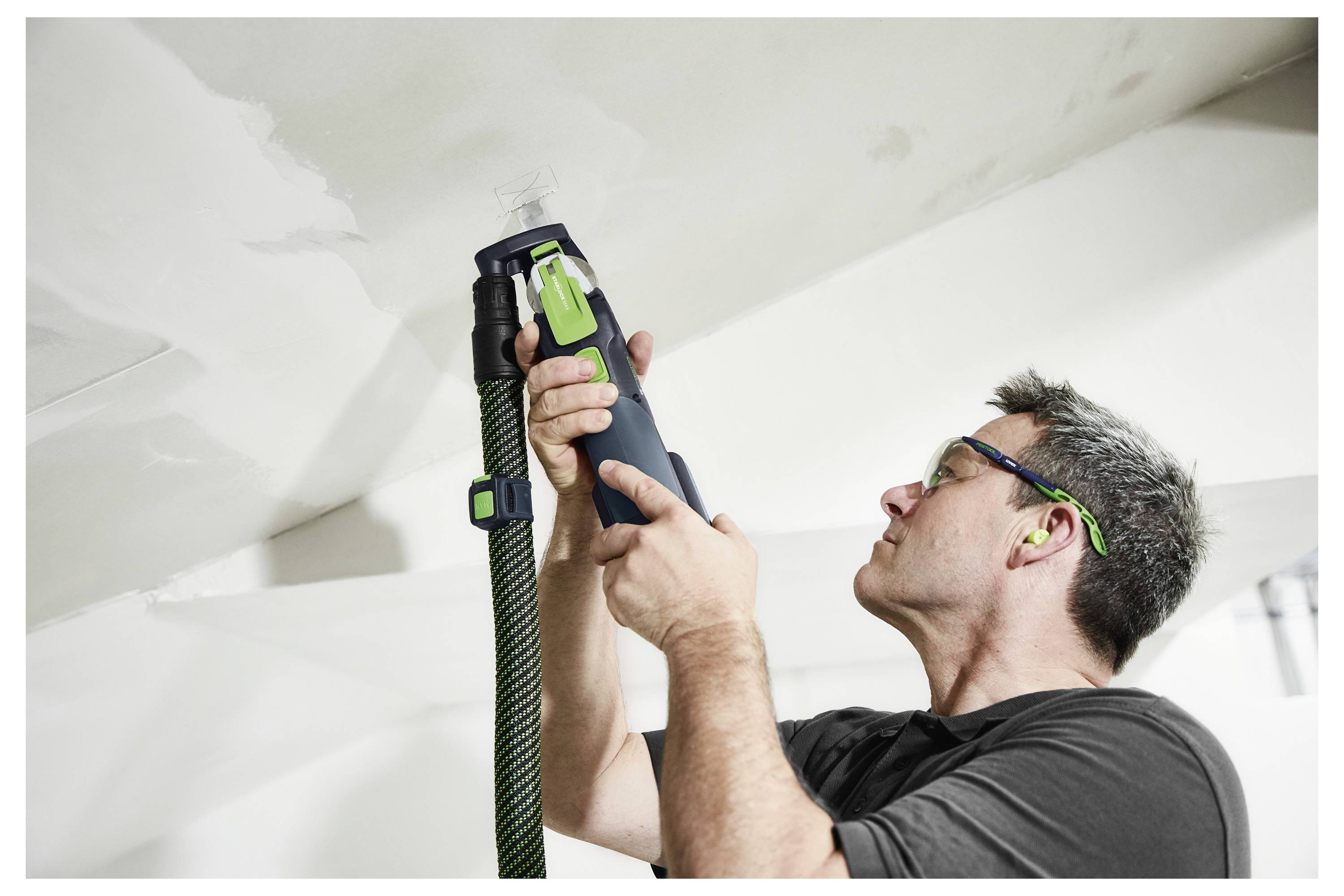 Osoba používající elektrické nářadí u bílé stěny, která má na sobě ochranné brýle. Zaměření je na nářadí a na povrch, na kterém se pracuje.