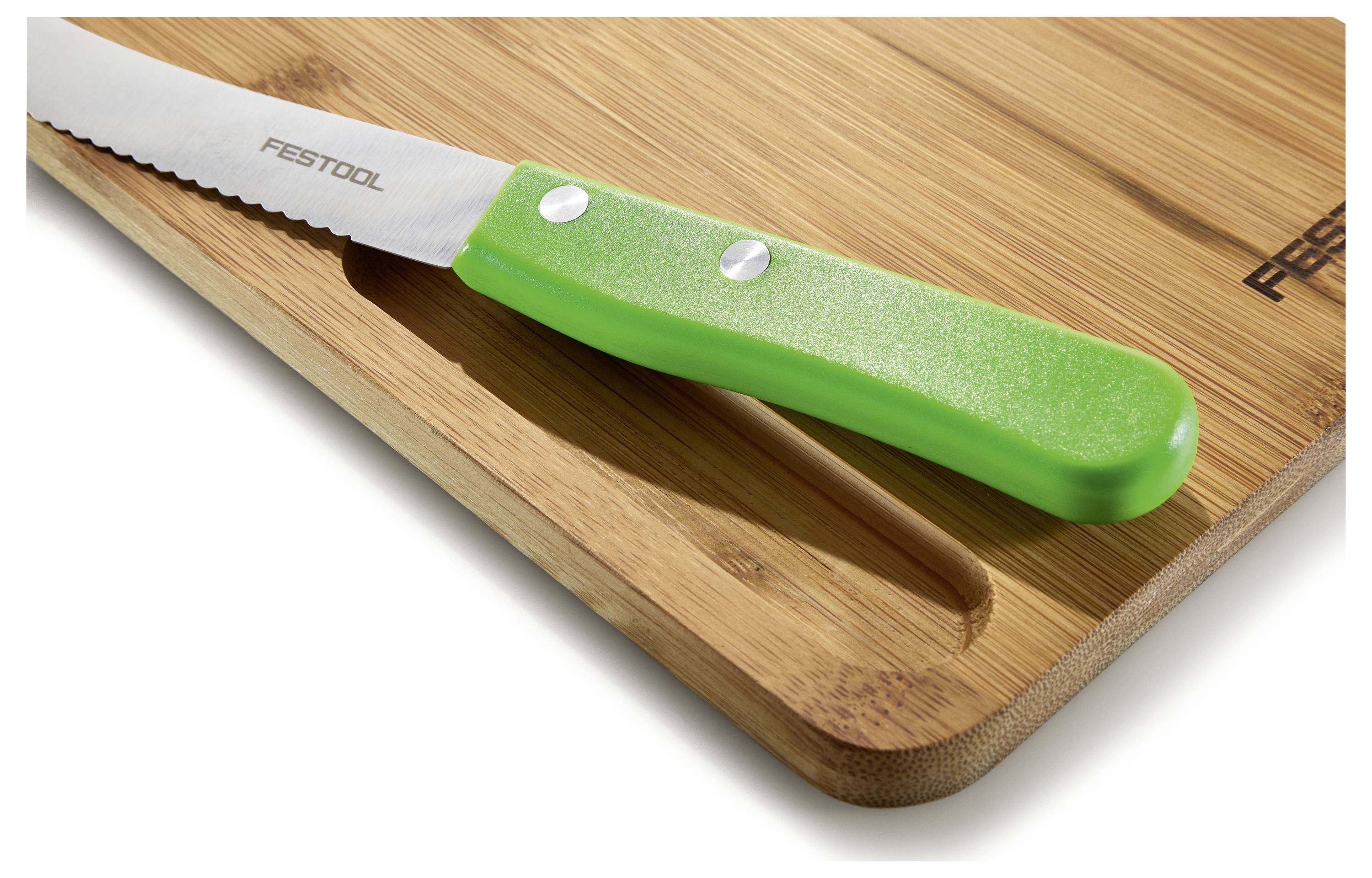 Vroubovaný nůž se zelenou rukojetí spočívá na bambusové kuchyňské desce s brandingem "Festool". Deska má drážku podél jedné hrany.