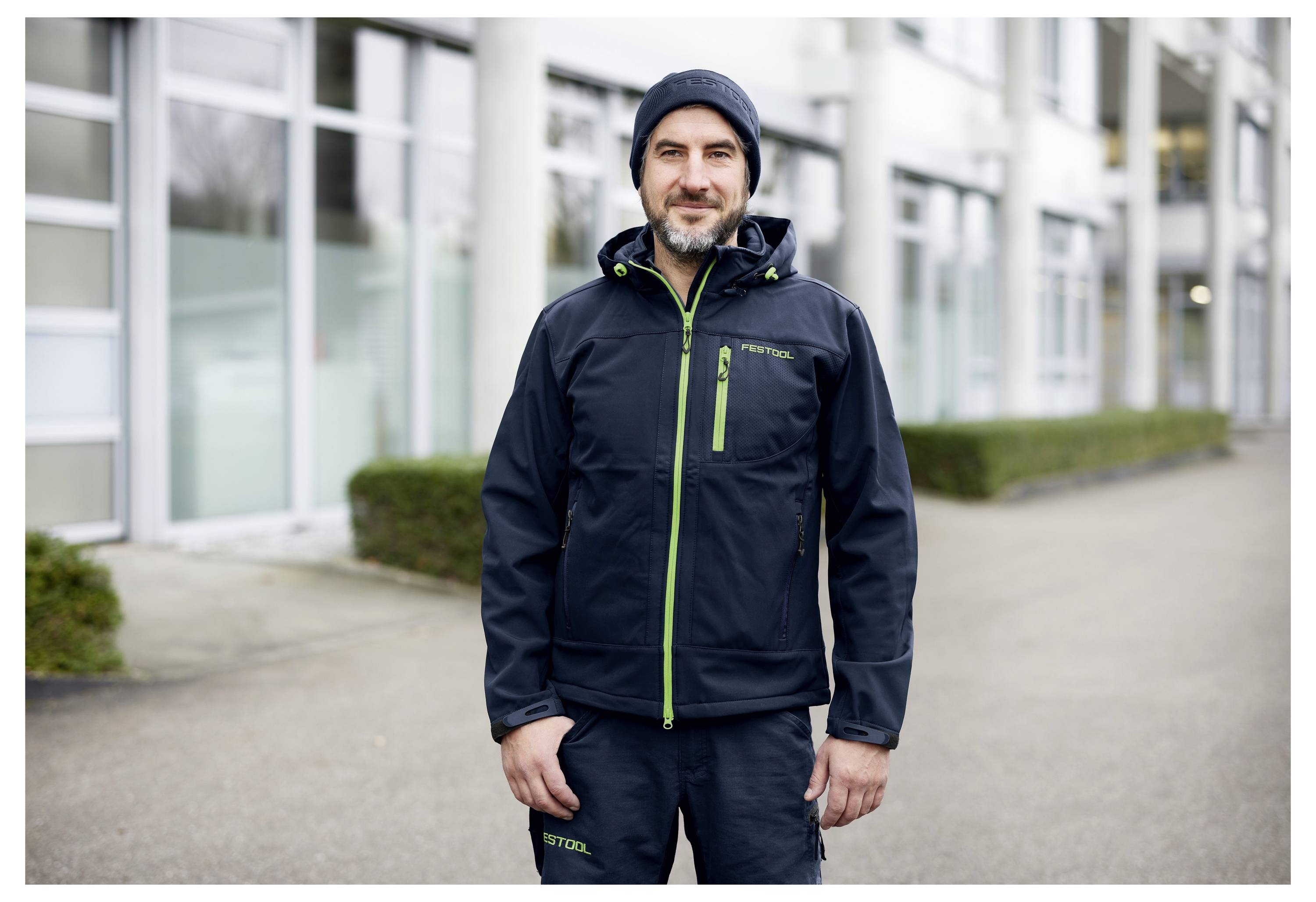 Muž v outdoorovém sportovním oblečení stojí s úsměvem, má na sobě čepici a bundu. Stojí venku před moderní budovou s velkými okny a sloupy.