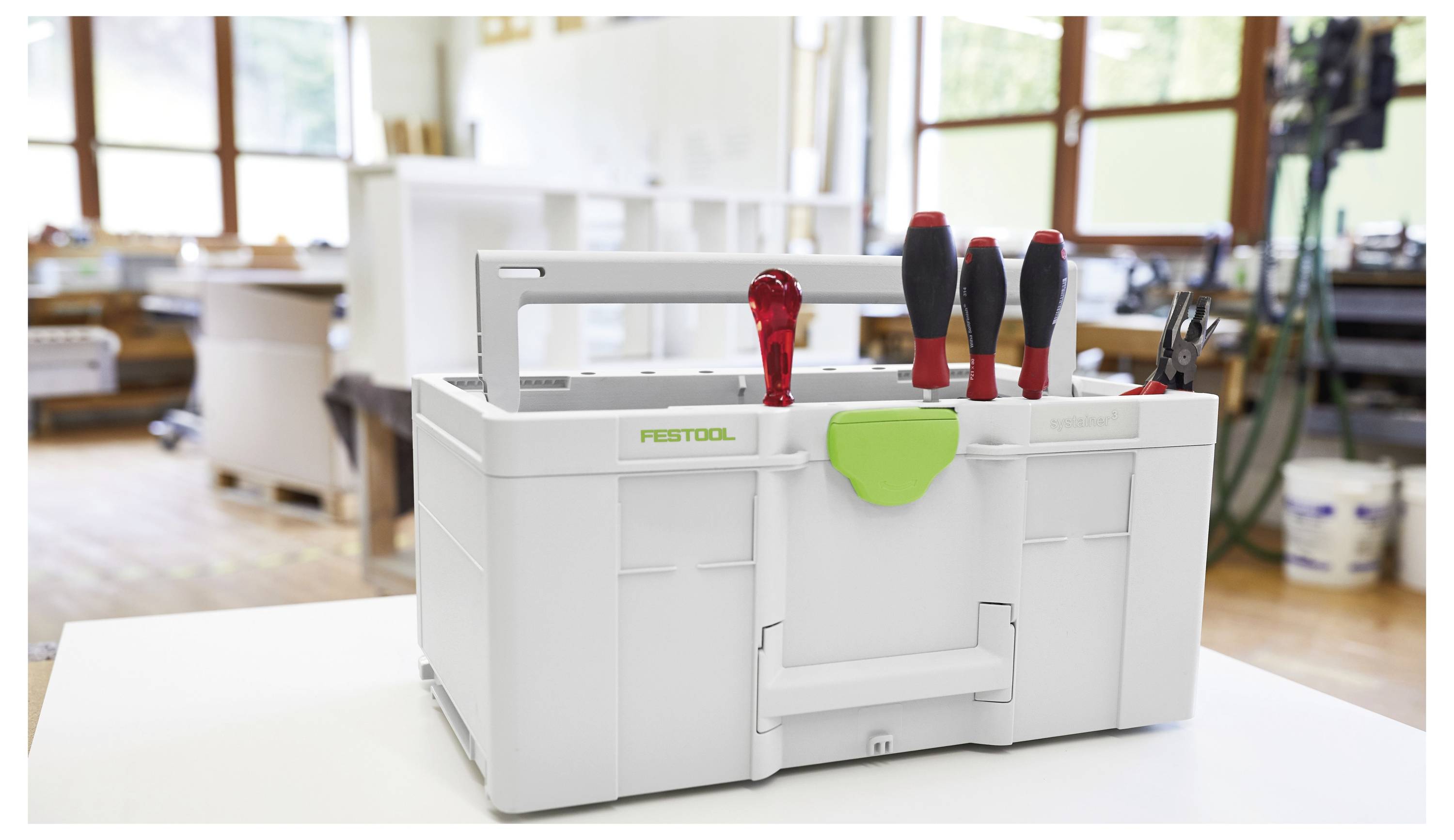 Bílý kufřík značky 'Festool' obsahuje několik šroubováků a kleští na pracovním stole v dílně, v pozadí jsou viditelné tesařské nářadí.