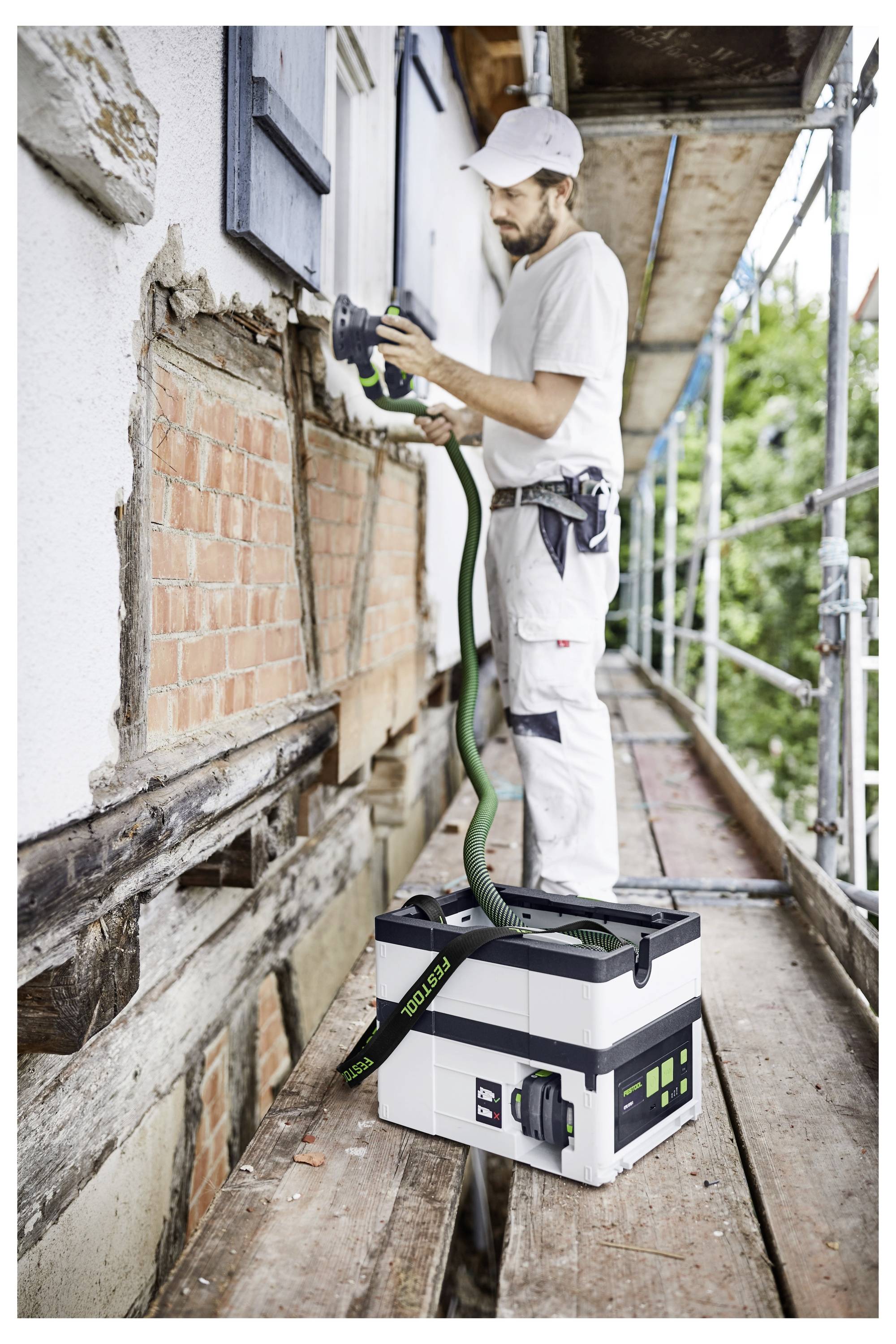 Pracovník v bílých montérkách používá elektrické nářadí na cihle na vnější straně domu, stojí na lešení s kufrem nářadí v blízkosti.