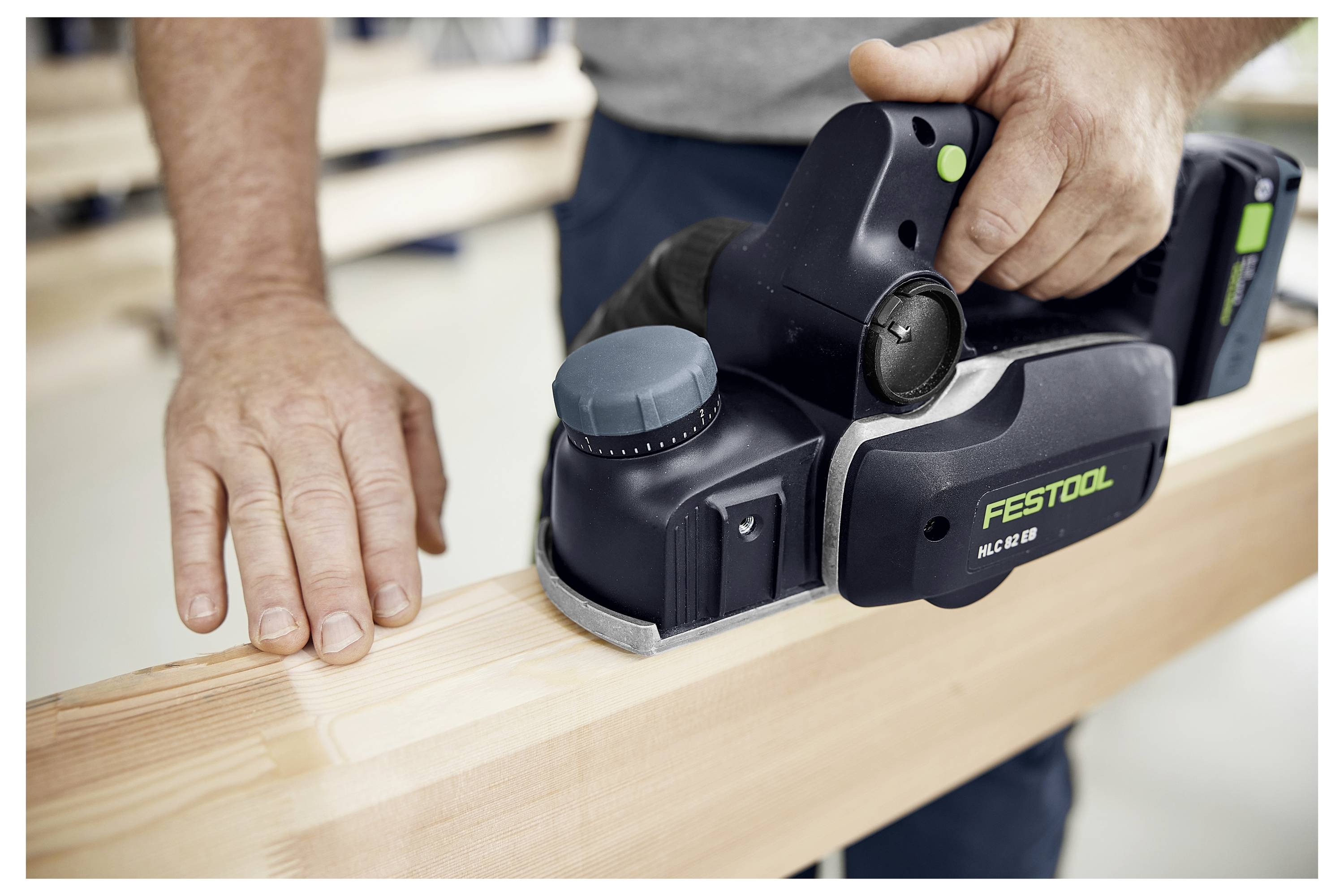 Osoba používající ruční hoblík Festool HG 82 EB na dřevěném trámě. Nástroj má černý a zelený design a ruka osoby vede dřevo.