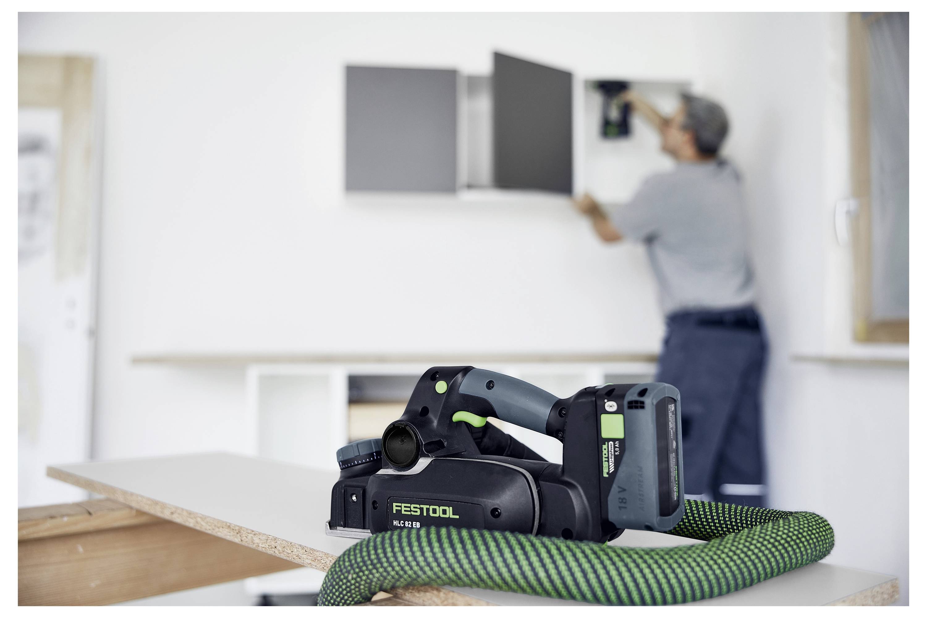 Muž instaluje skříňky v místnosti. Zaměření je na elektrickém stroji Festool umístěném na pracovním stole v popředí s připojenou hadicí.