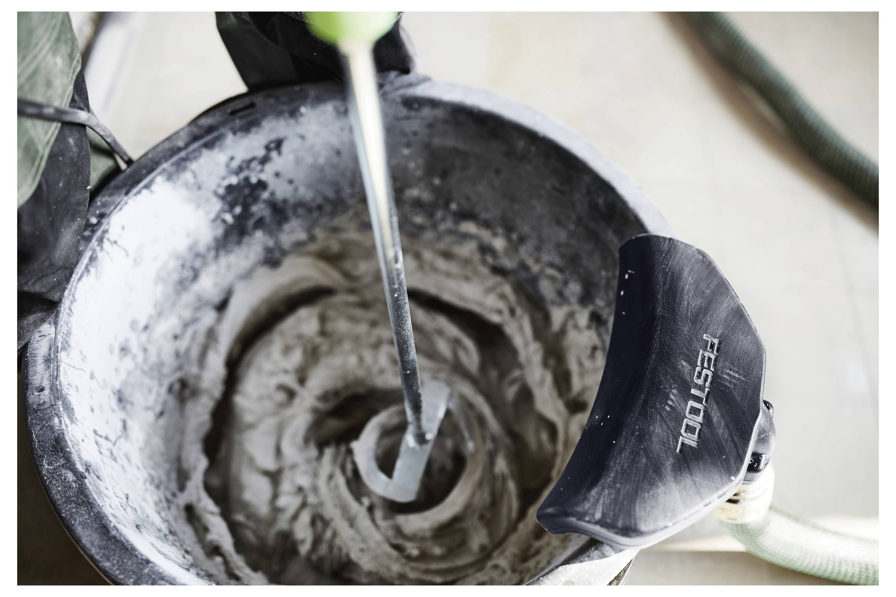 Míchací nástroj míchá cement v kbelíku, což naznačuje probíhající stavební nebo renovační práce. Název značky nástroje je částečně viditelný.