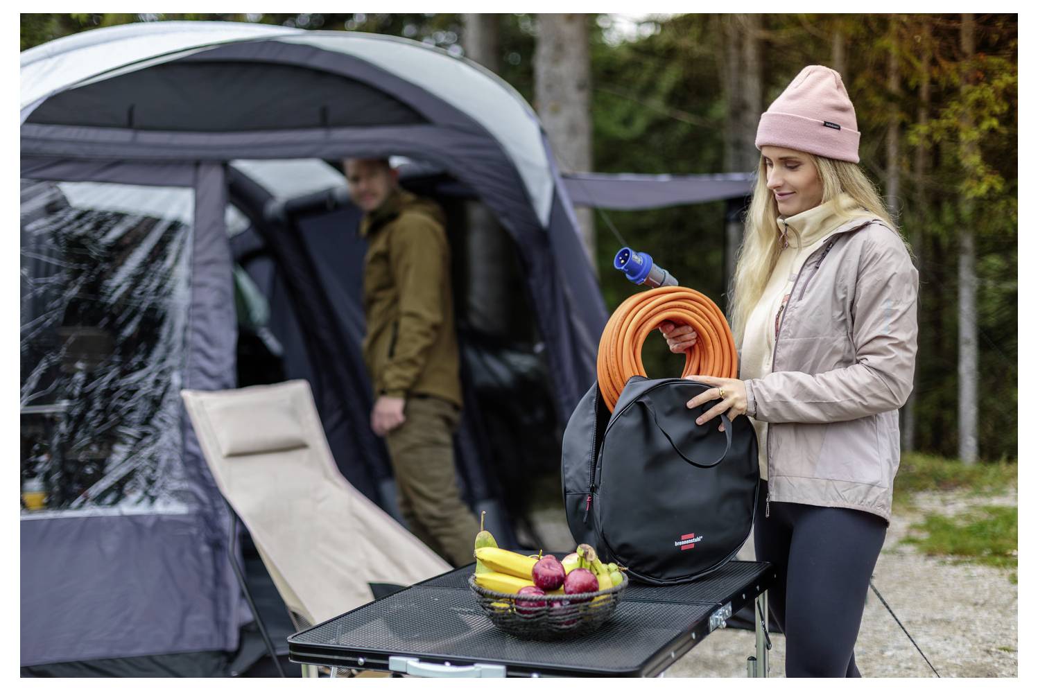 Žena balící camping vybavení vedle stanu, drží batoh se svitnutou oranžovou hadicí; piknikový stůl s košem ovoce v popředí.