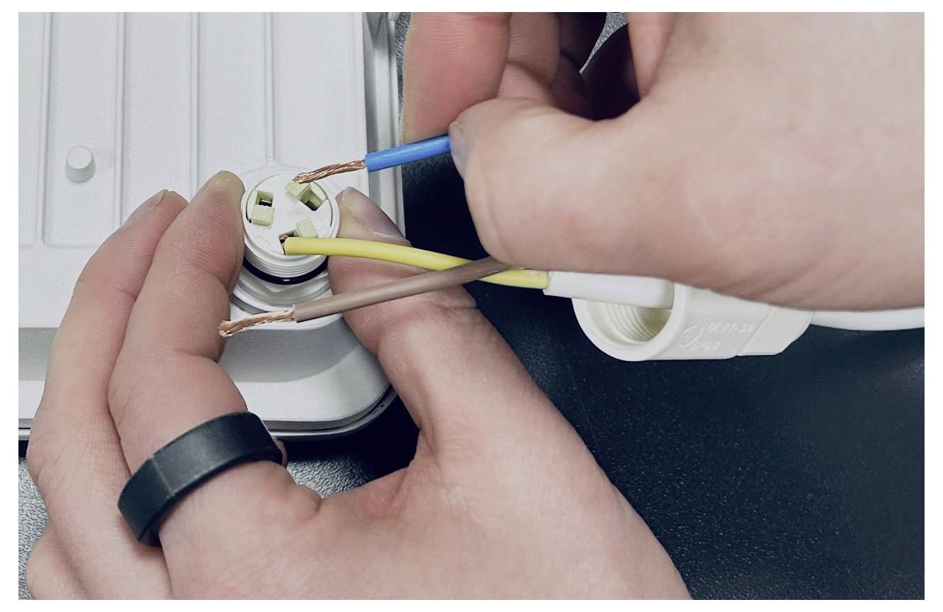 Ruce zapojují bílou elektrickou zásuvku a připojují modrý, hnědý a žluto-zelený kabel do objímky.