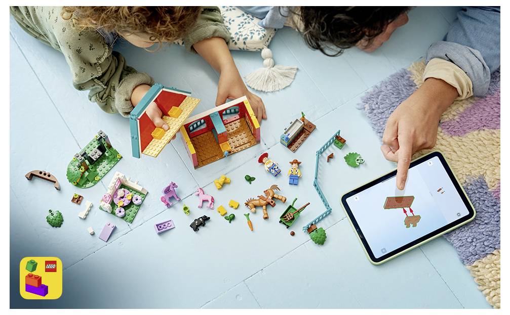 Dva lidé stavějí dům z LEGA a hrají si s rozházenými kostičkami, zatímco ukazují na tablet zobrazující hračkařskou loď na obrazovce.
