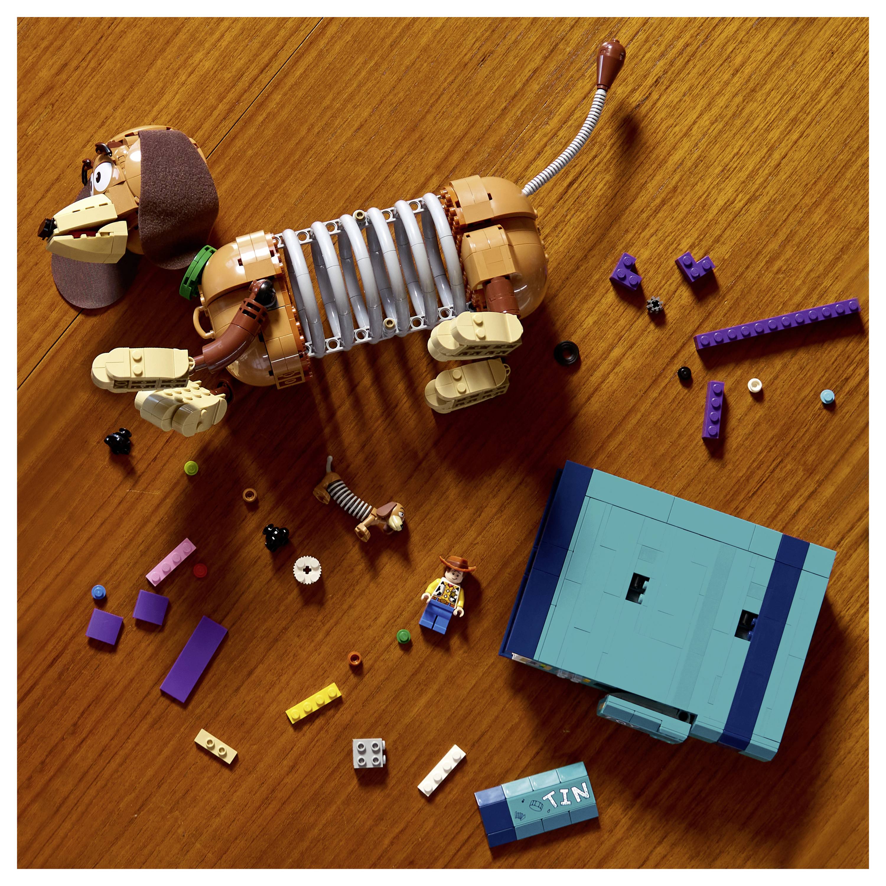Pohled shora na kostru psa z LEGO vedle modré LEGO krabice, rozptýlené LEGO kostičky a malá minifigurka na dřevěném stole.