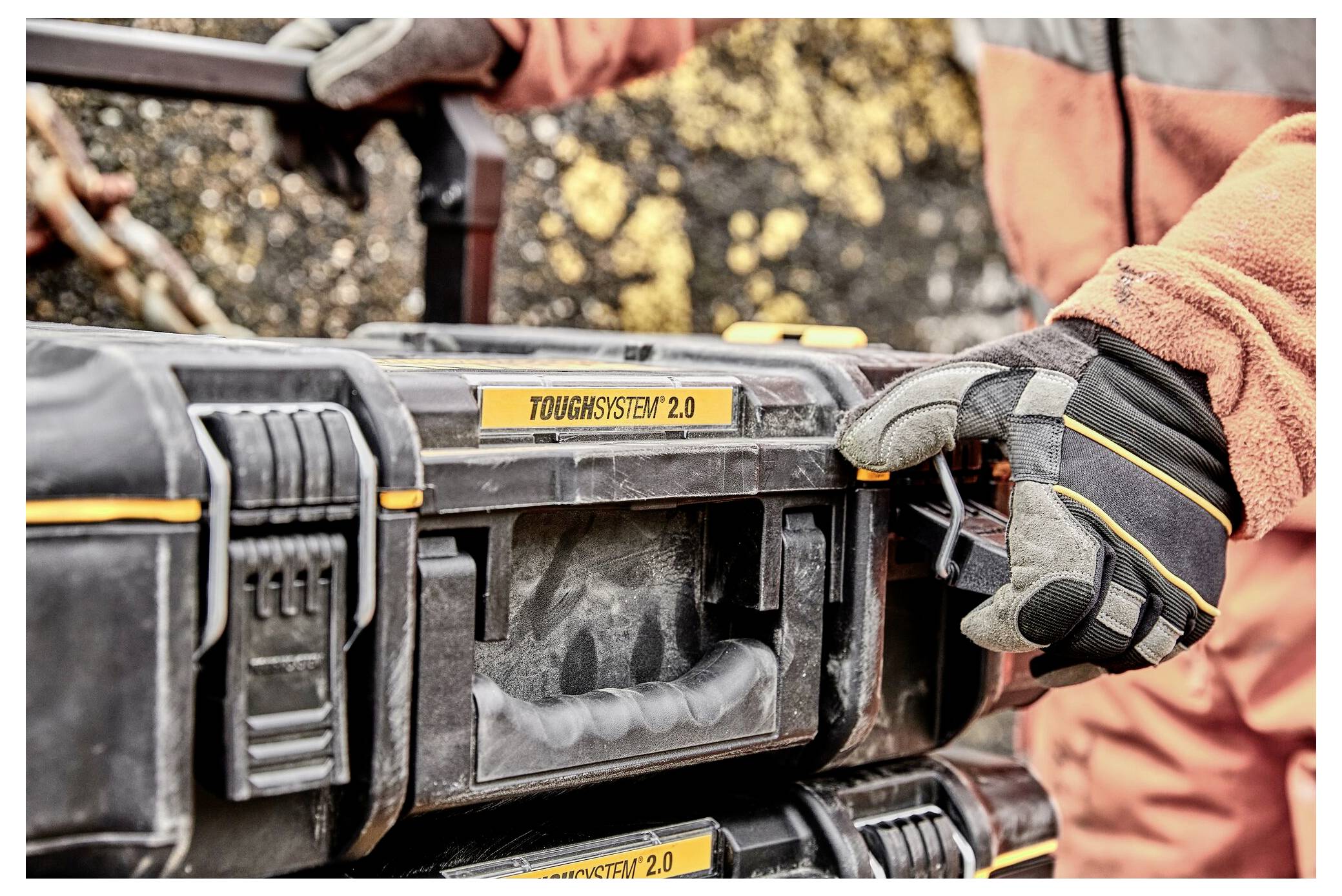 Osoba v rukavicích drží a otevírá černožlutou skříňku s nástroji označenou "Toughsystem 2.0" venku, což naznačuje pracovní nebo opravárenské prostředí.