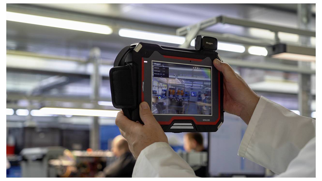 Osoba v laboratorním plášti drží tabletové zařízení zobrazující digitální rozhraní, které zachycuje živý obrázek interiéru výrobního závodu.