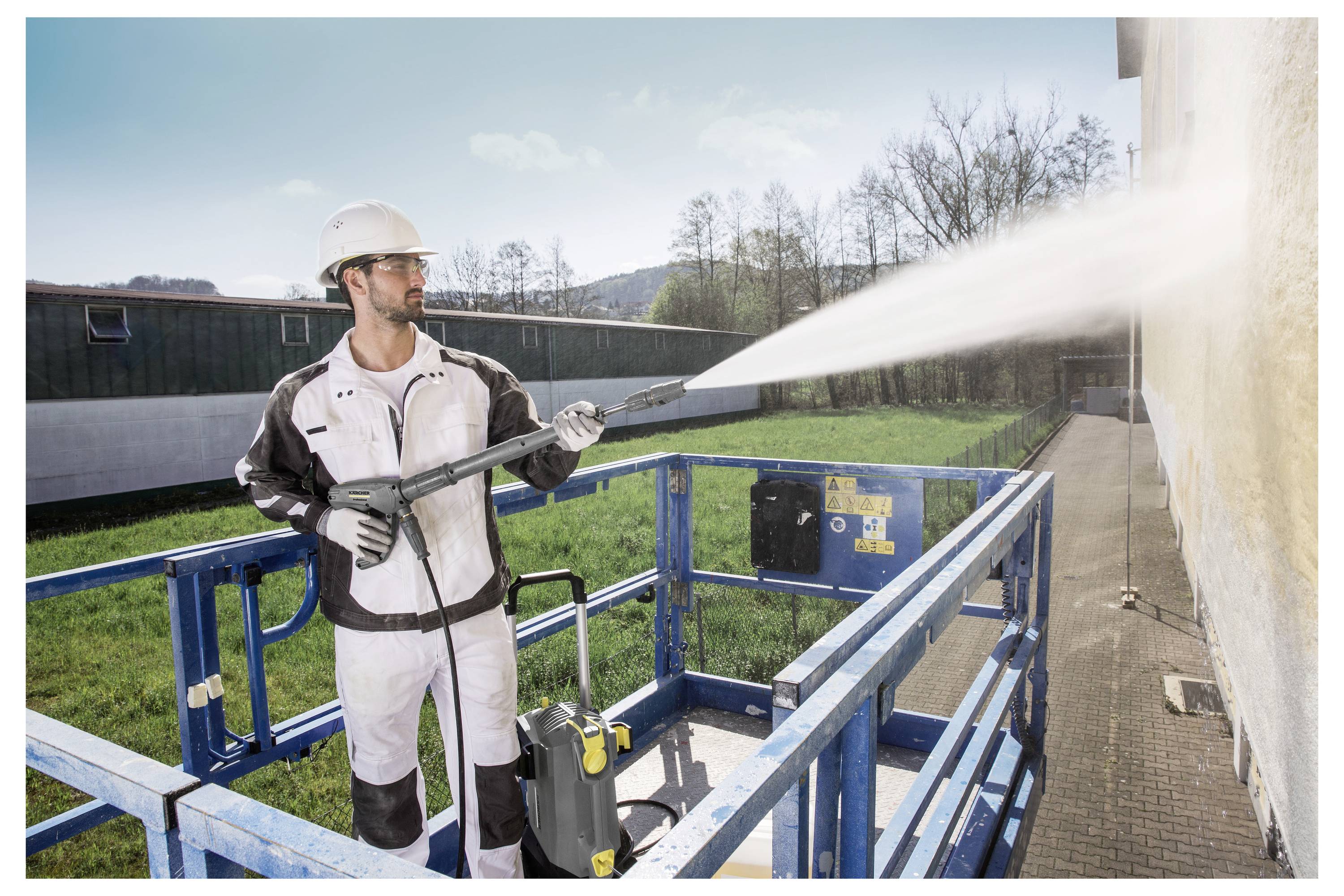 Dělník v ochranném oděvu používá vysokotlaký čistič k čištění vnějšku budovy a stojí na modré plošině zvedáku venku.