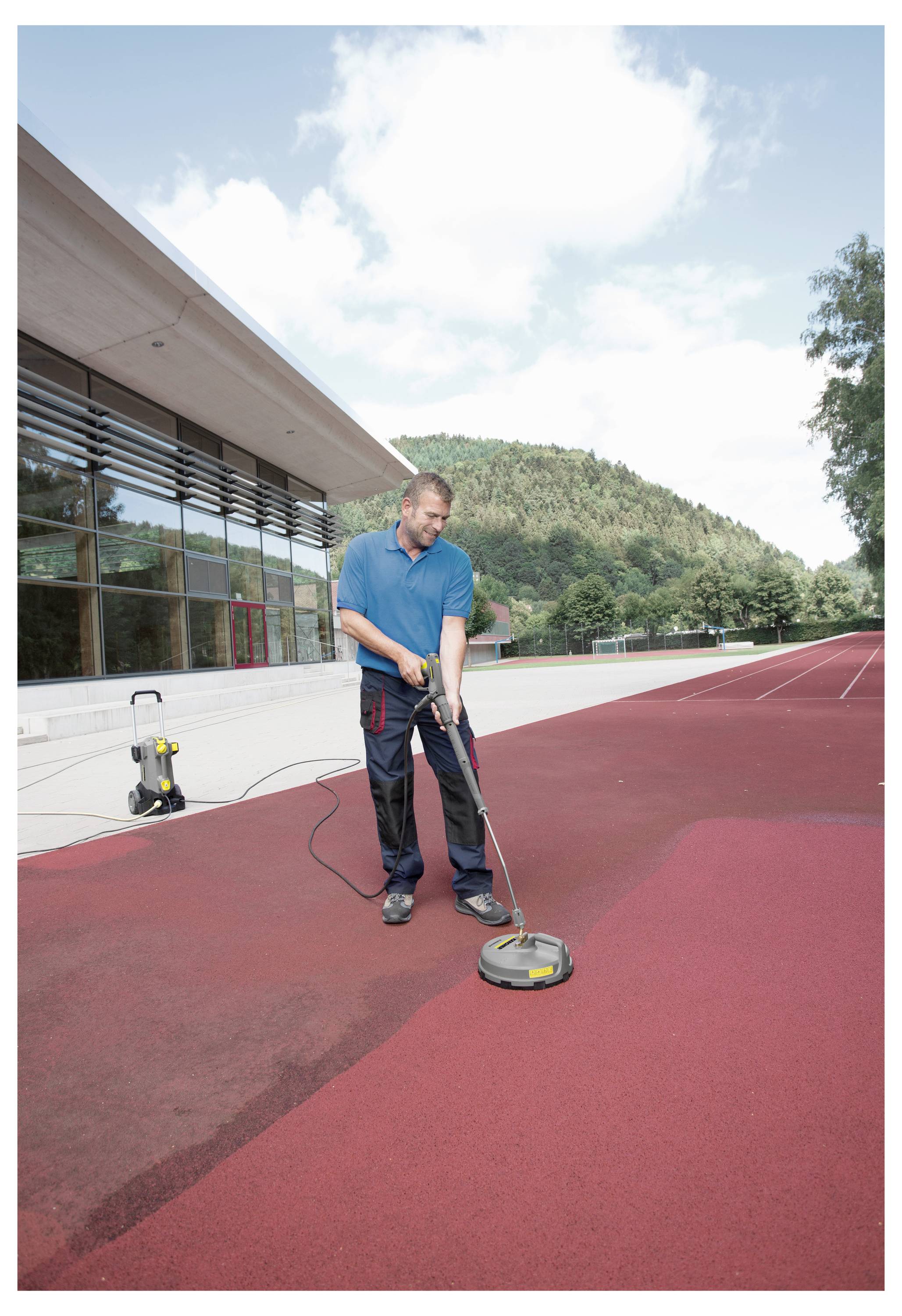 Osoba čistící červenou venkovní atletickou dráhu vysokotlakým čističem, vedle moderní budovy a stromů v pozadí.